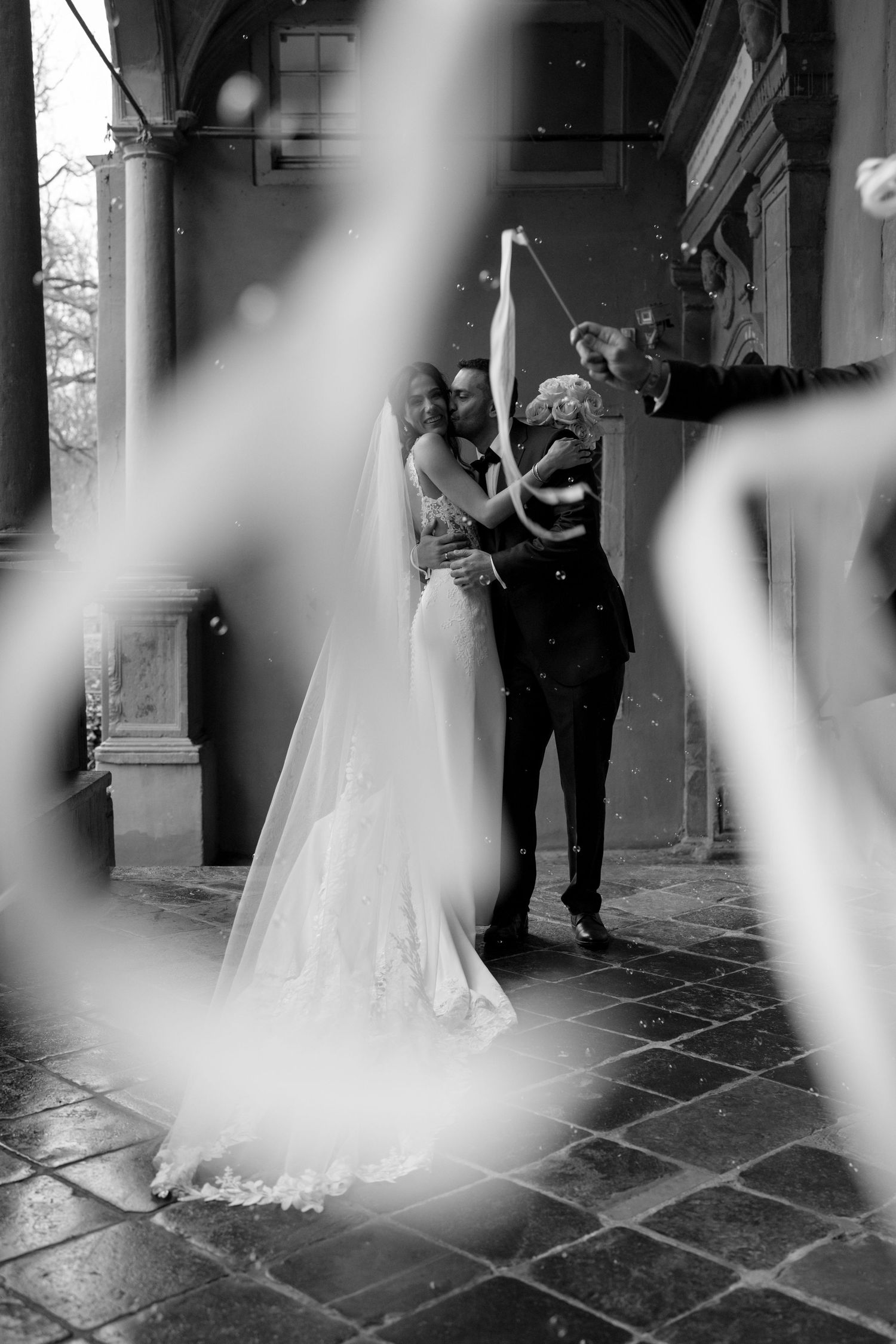 Bride and groom celebrate with confetti under historic archway captured through artistic foreground blur in black and white.