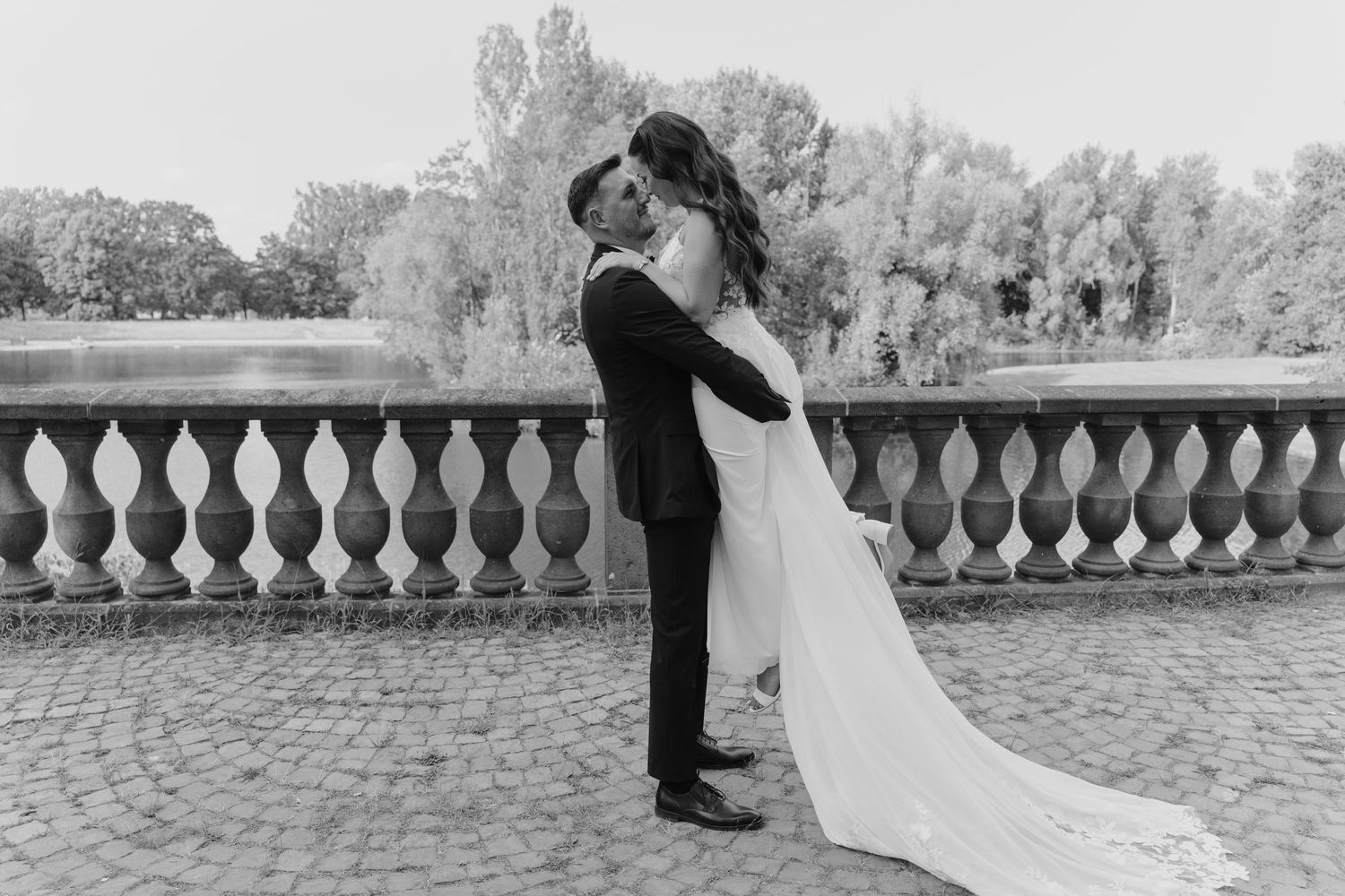 Bride and groom embrace near ornate stone balustrade in elegant black and white outdoor wedding portrait.