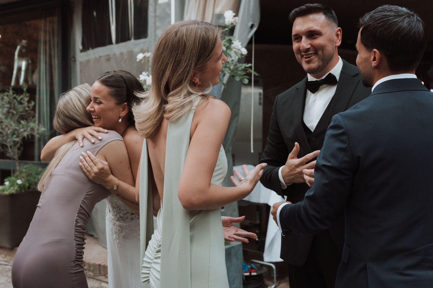 Guests greeting each other at a wedding reception, with women hugging and men shaking hands outdoors.