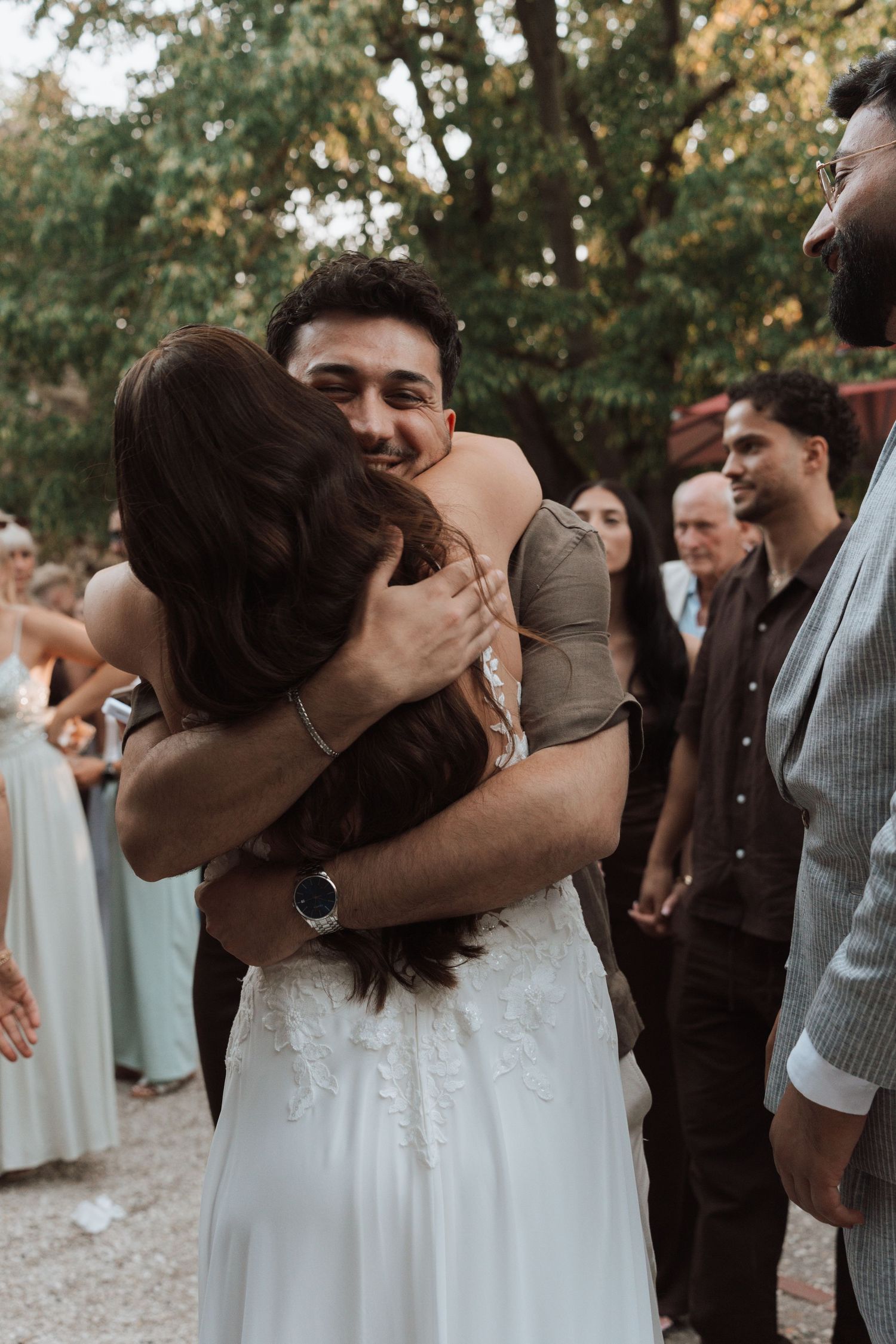 Smiling groom embracing bride in lace wedding dress while guests celebrate in background.