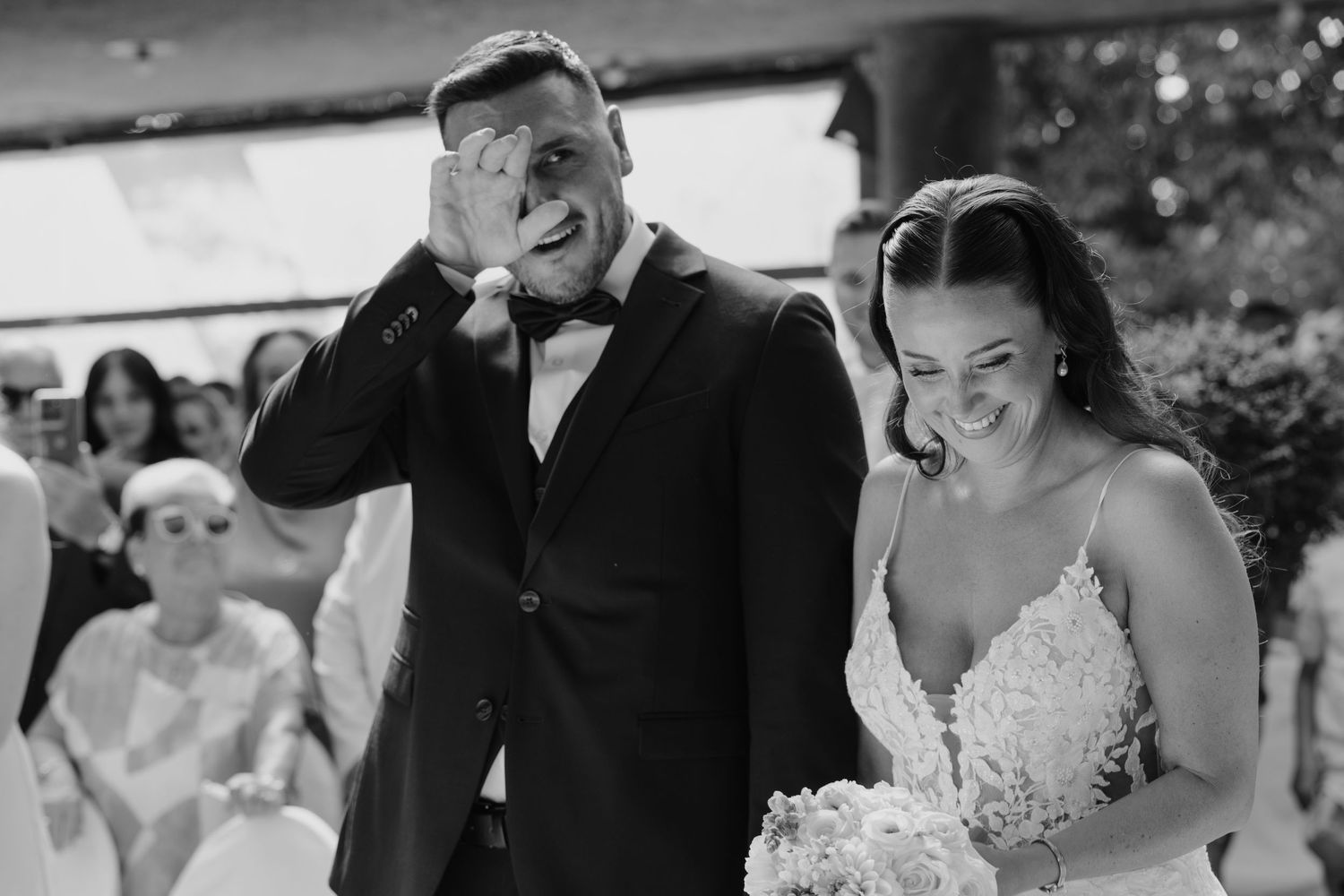 Emotional groom wipes tears while smiling bride holds bouquet during outdoor wedding ceremony.