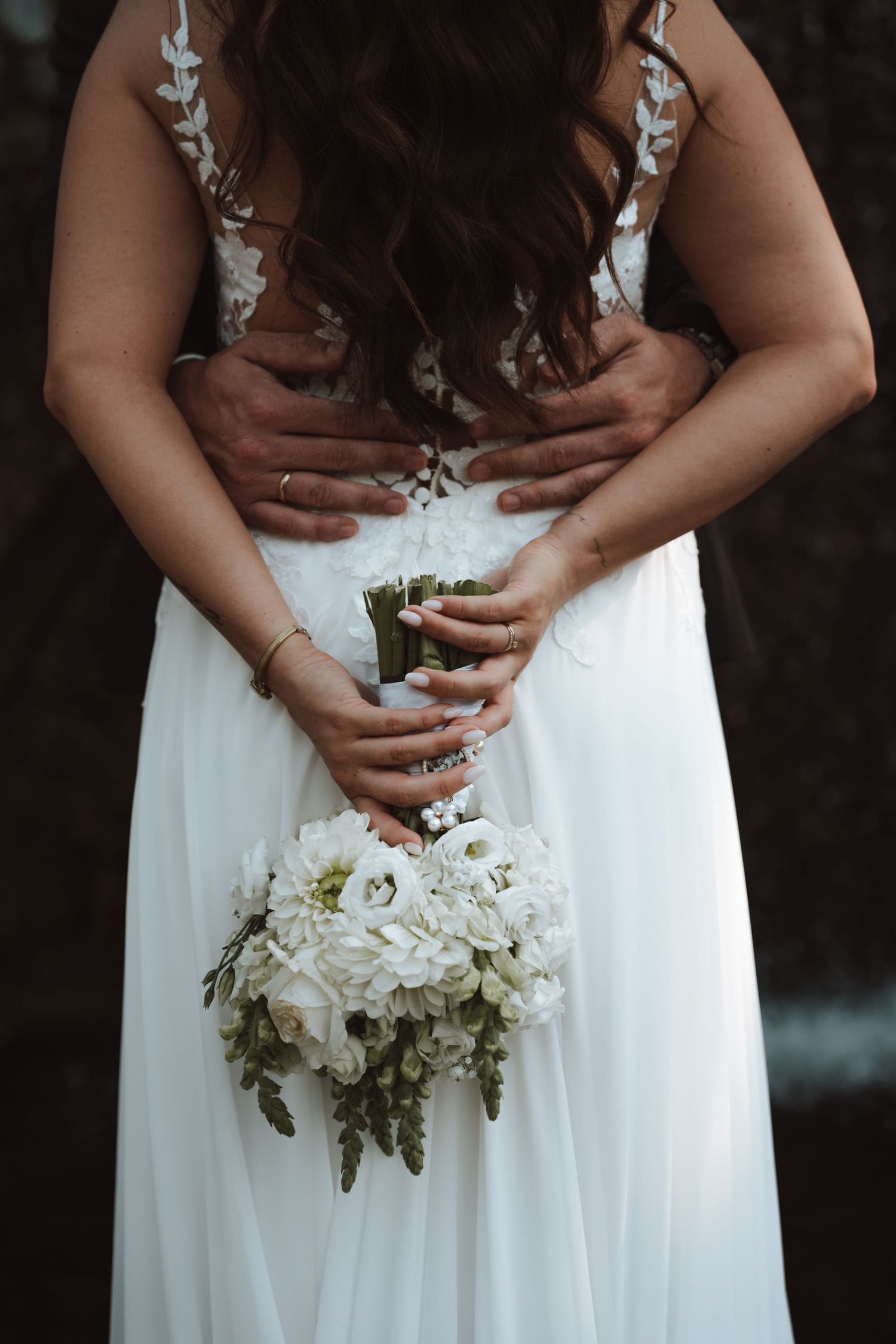 Bride in white lace gown holds bouquet of white flowers while groom embraces her from behind.