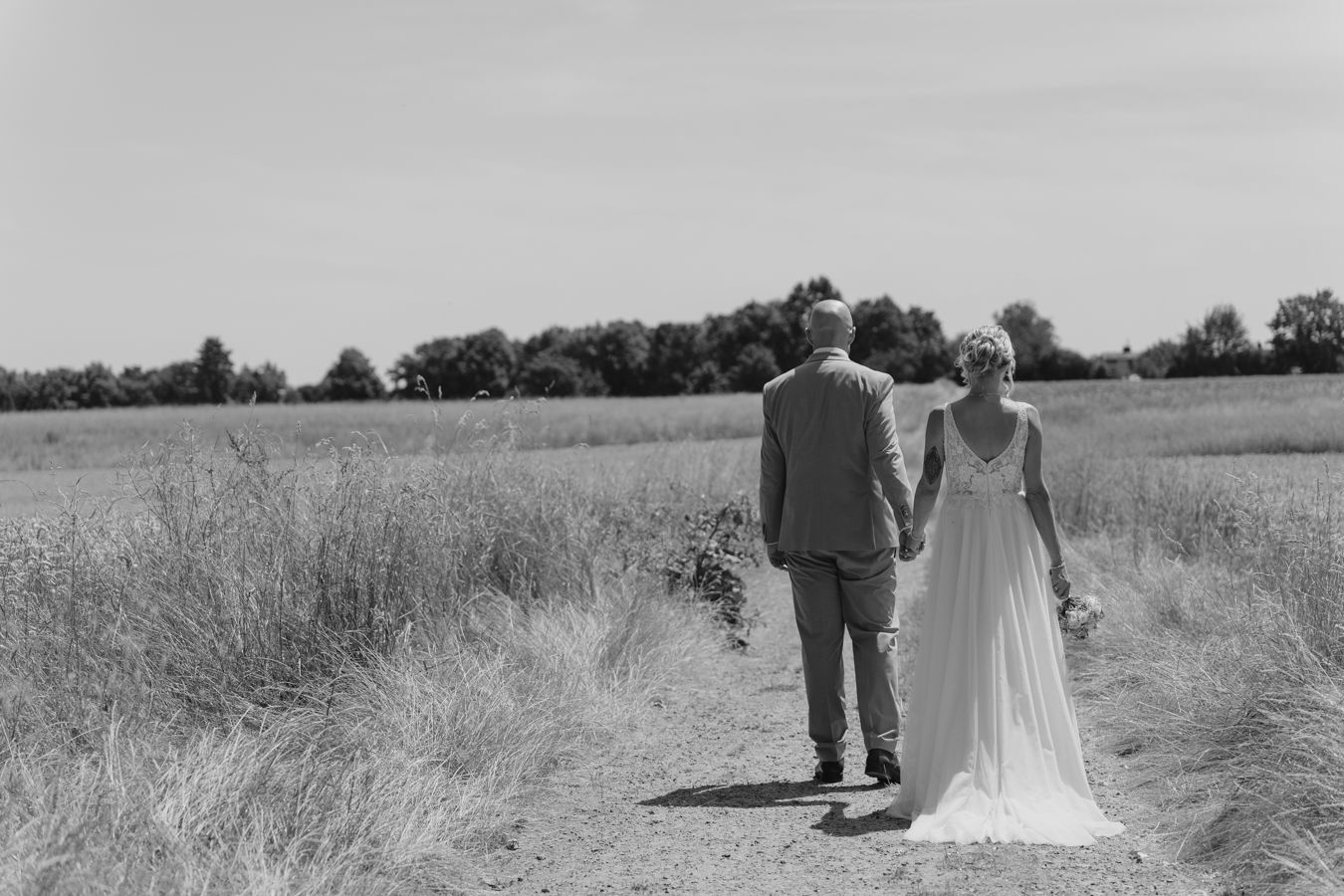 Bride and groom walk hand in hand along rural path through golden fields.