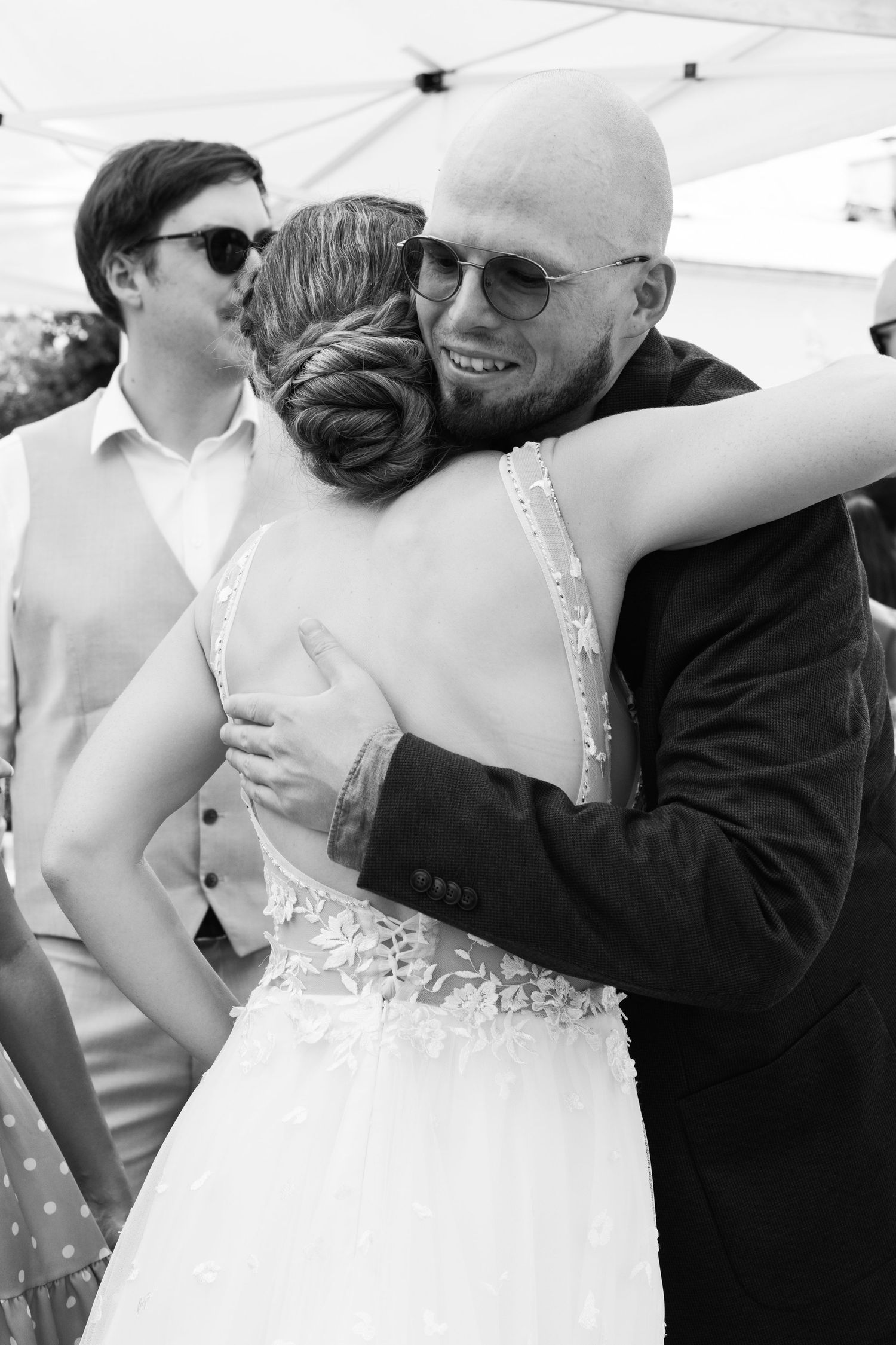 Bald man in sunglasses shares warm embrace with bride in lace wedding gown outdoors.