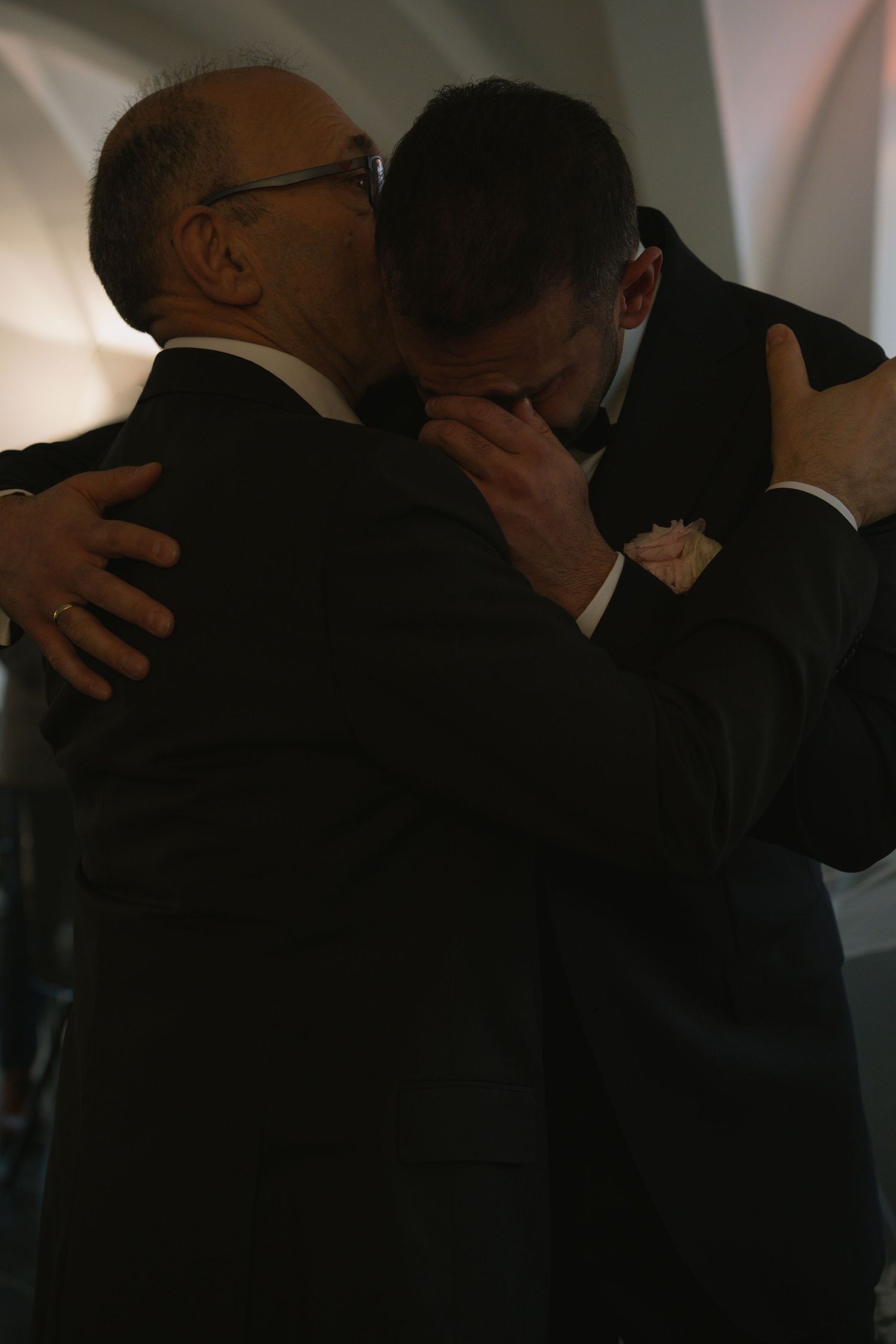 Two men in tuxedos share an emotional embrace in dimly lit wedding venue.