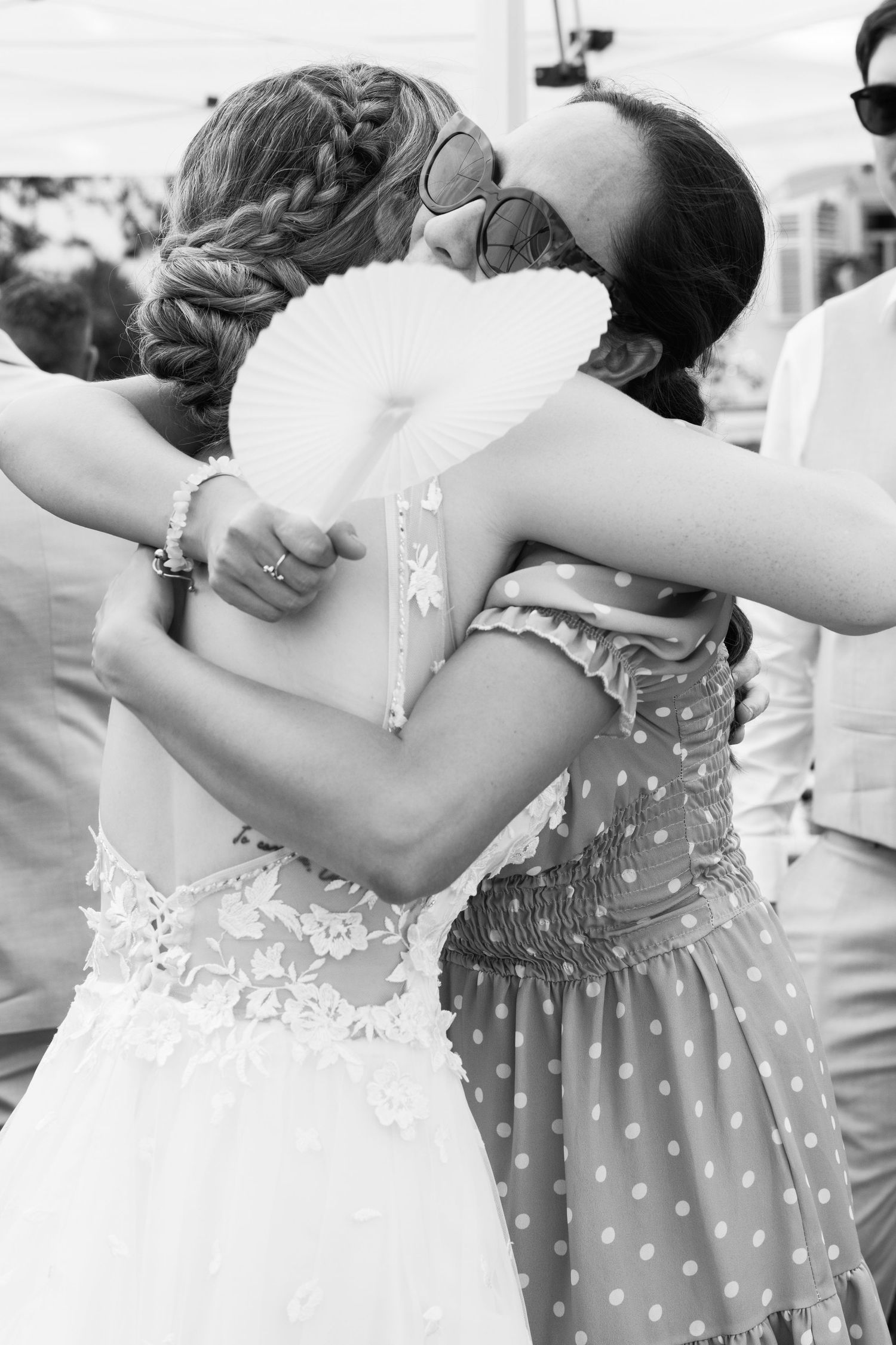 Black and white photo of bride in lace wedding dress sharing warm embrace with guest holding paper fan.