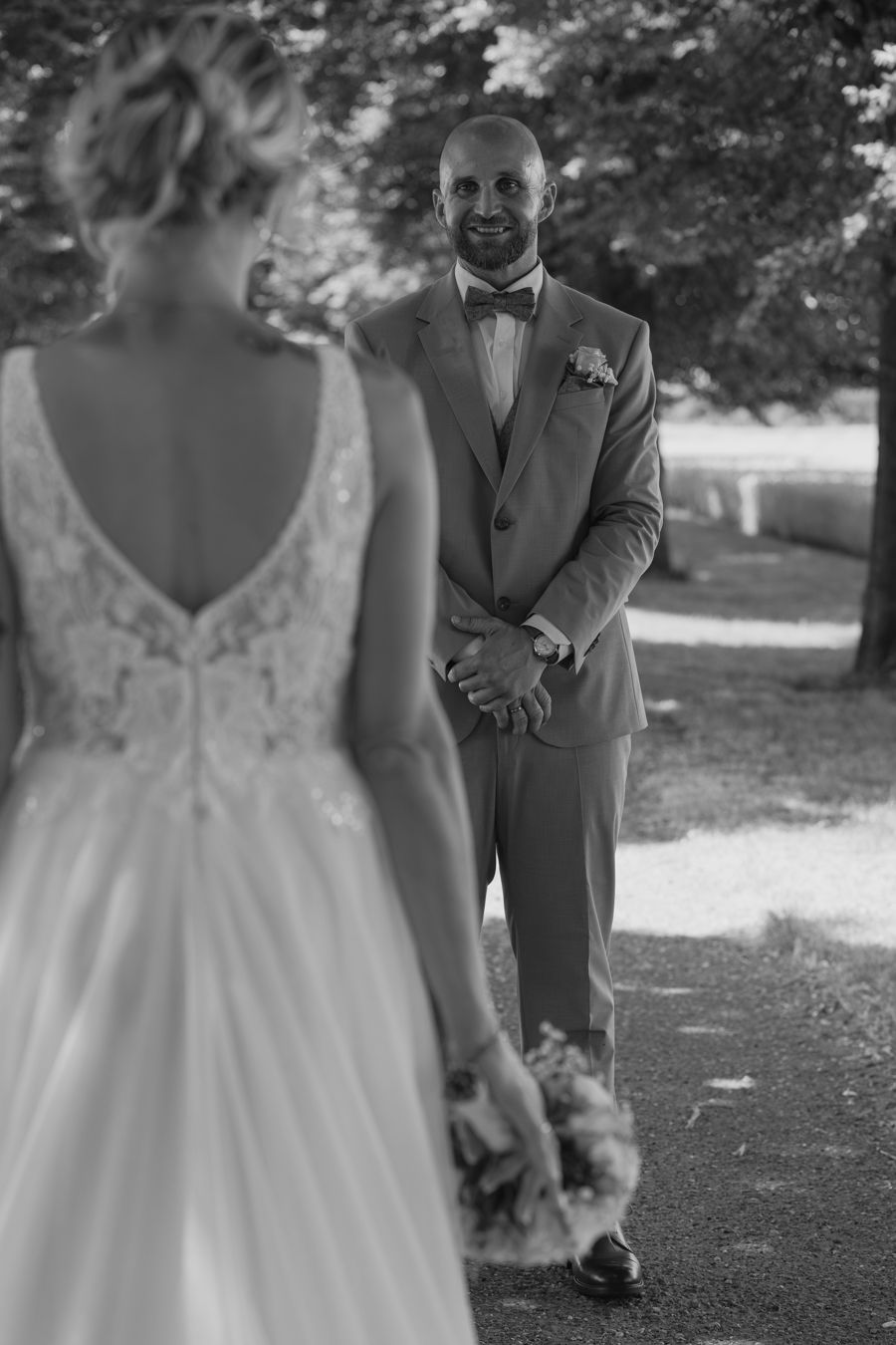 Groom in gray suit smiles at bride during wedding first look on outdoor path.