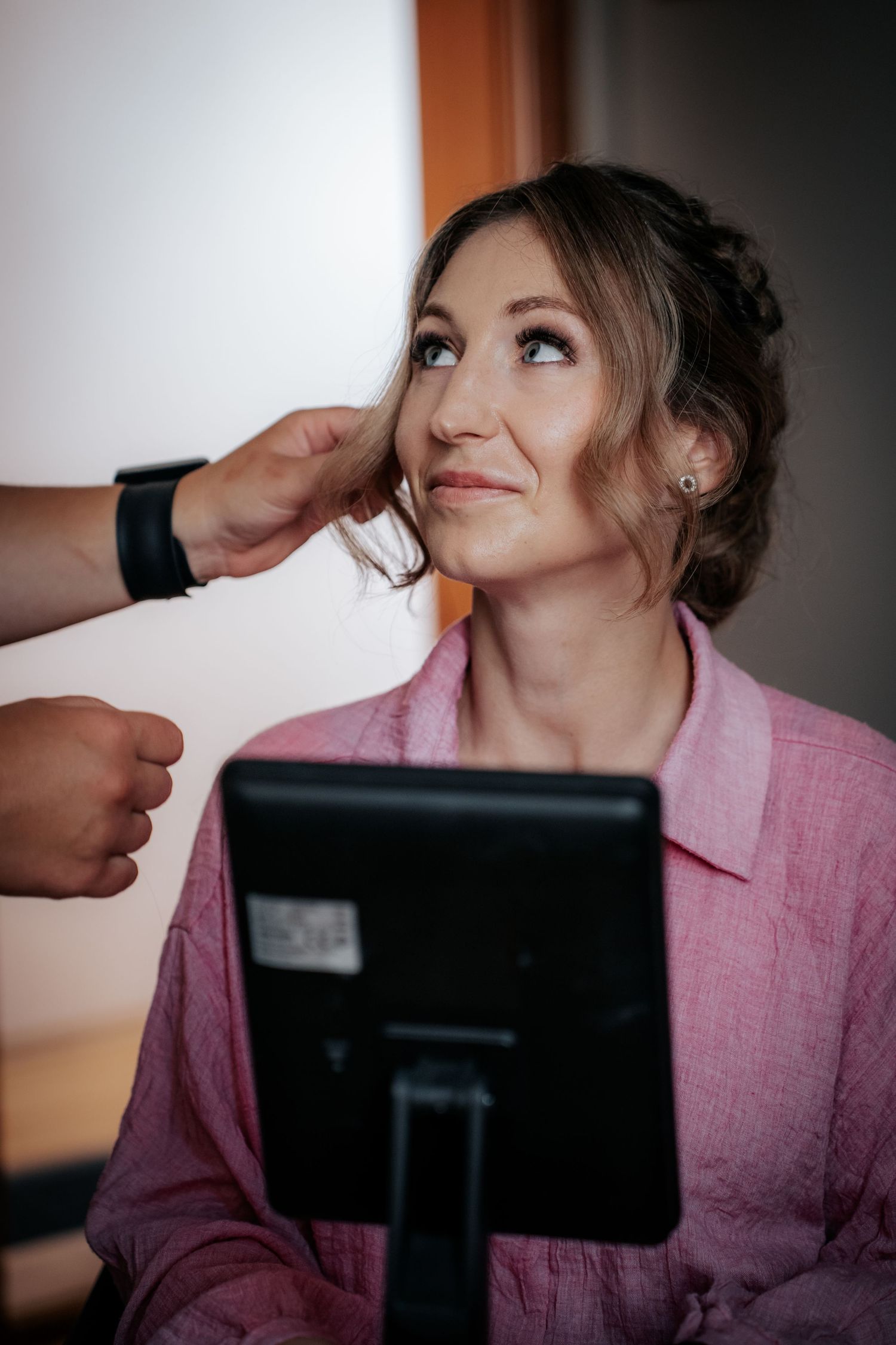 Bride getting hair and makeup done while sitting in front of a mirror in a pink robe.
