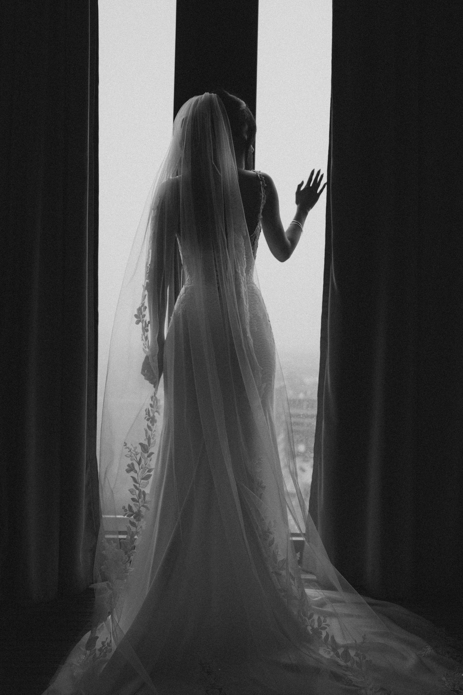 Black and white silhouette of bride in fitted gown and cathedral veil standing at large window.