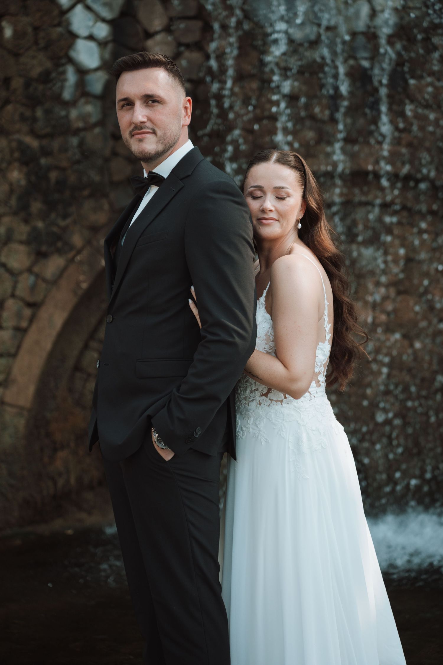 Bride leans against groom's back near stone waterfall feature in romantic wedding portrait outdoors.