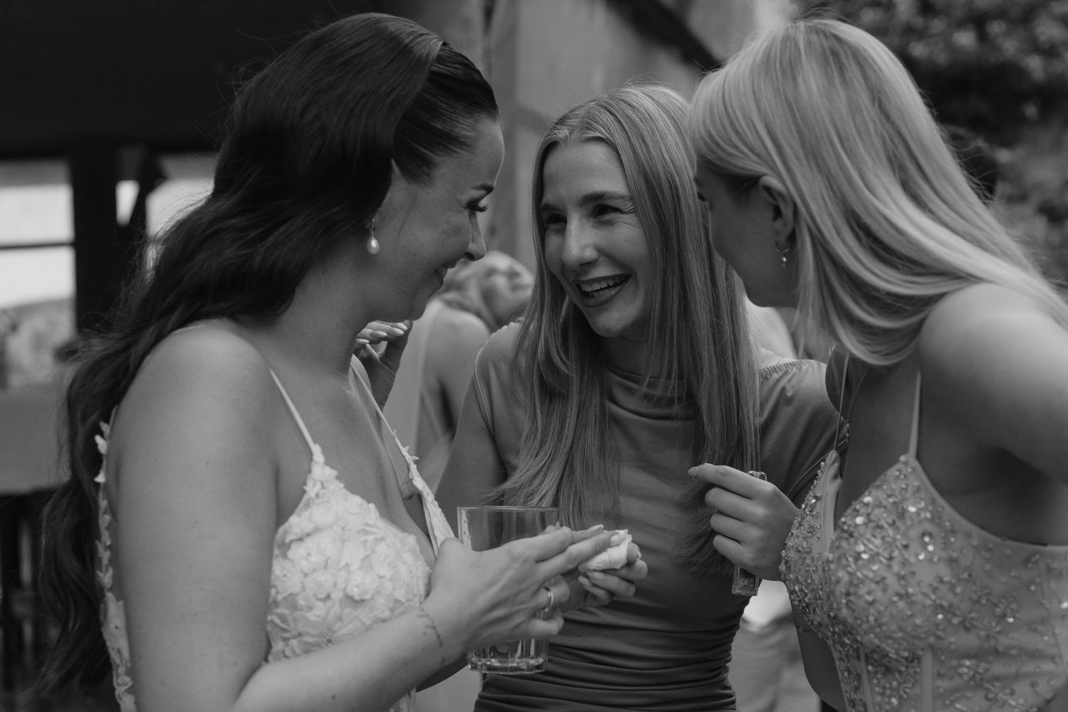 Three women in elegant dresses laughing together at outdoor wedding reception in black and white.