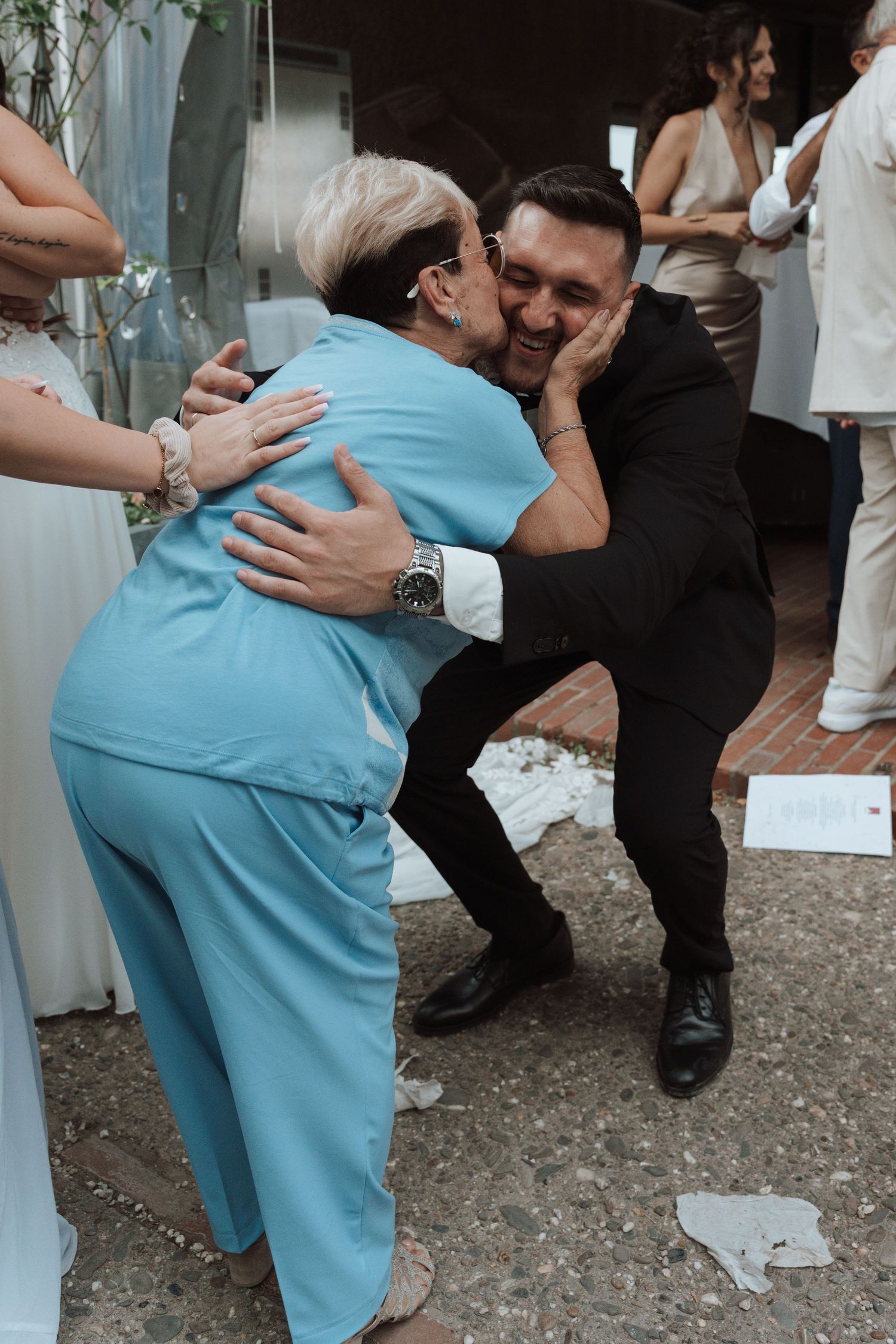 Elderly woman in blue outfit kissing groom on cheek during warm wedding congratulations moment.