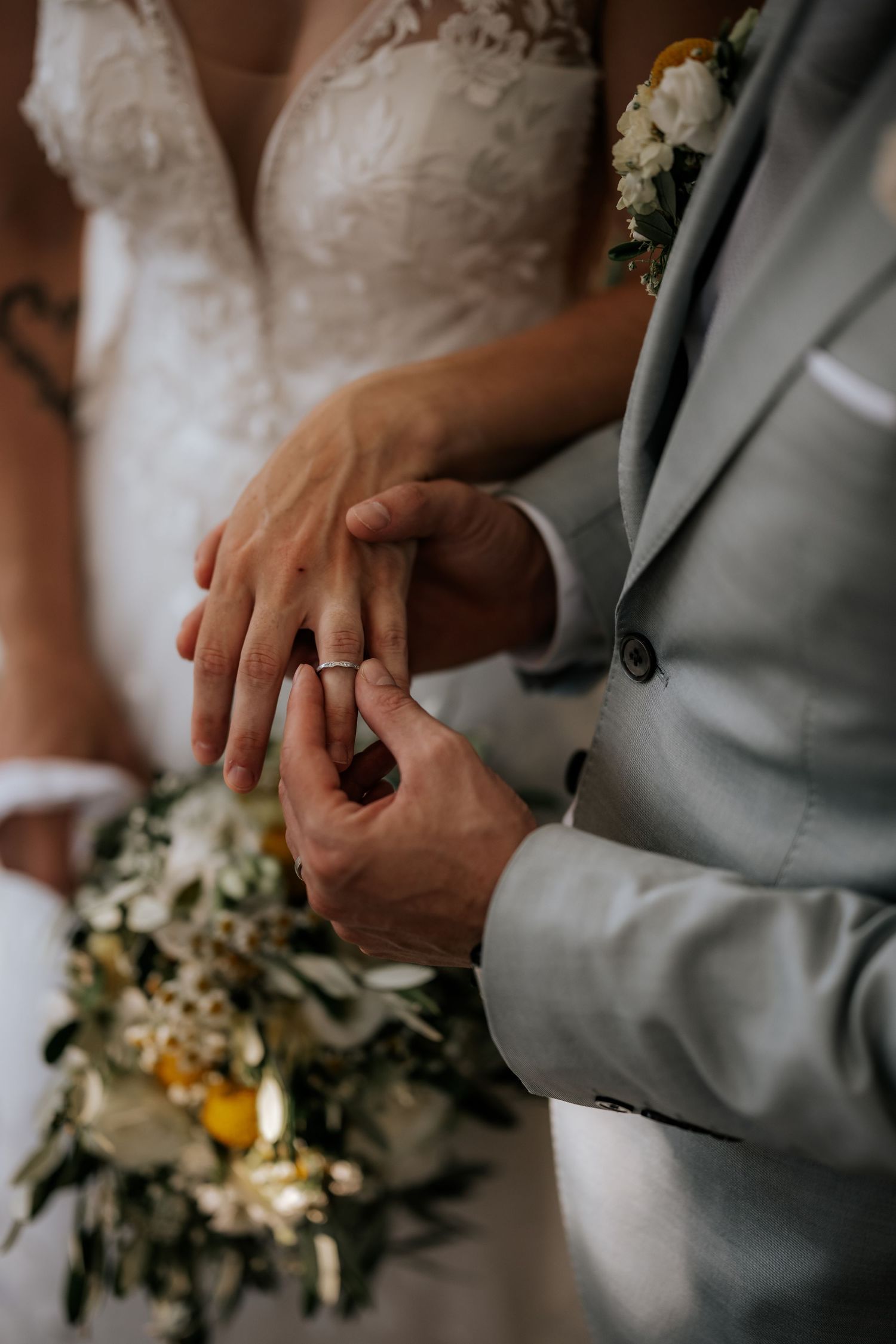 Groom placing wedding ring on bride's finger during ceremony with floral bouquet.