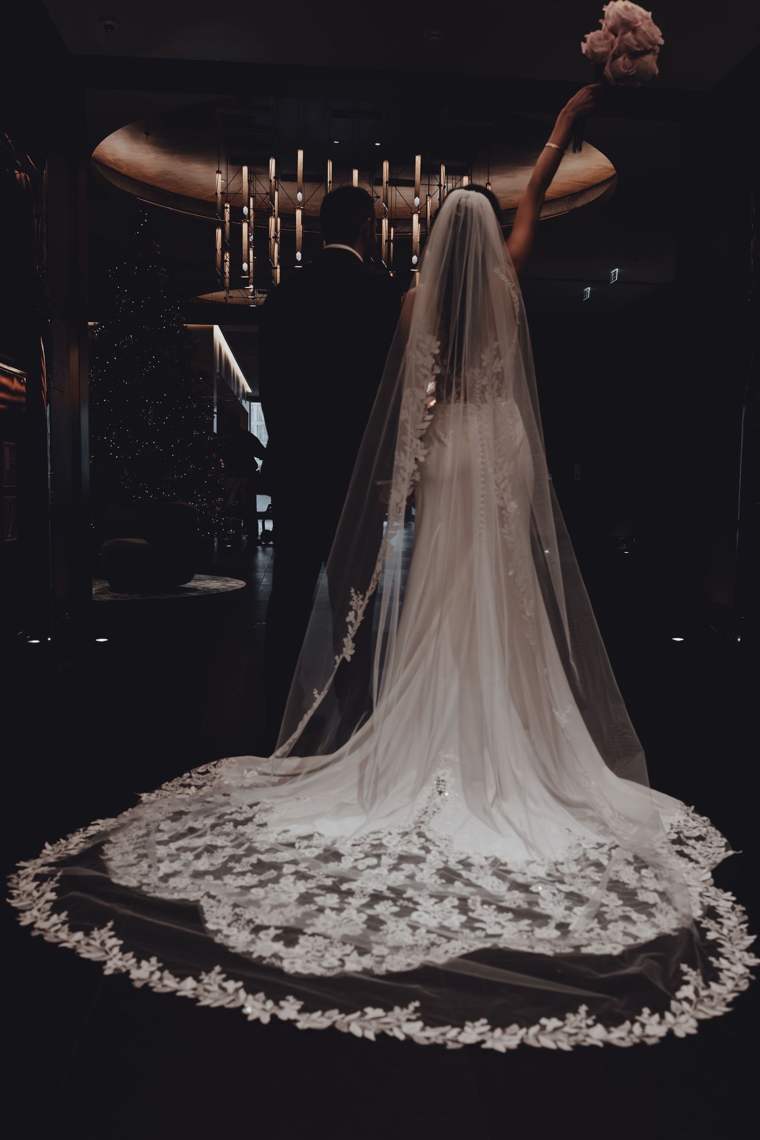 Bride in ornate cathedral-length lace veil stands with groom before candlelit altar in dramatic dark wedding photo.