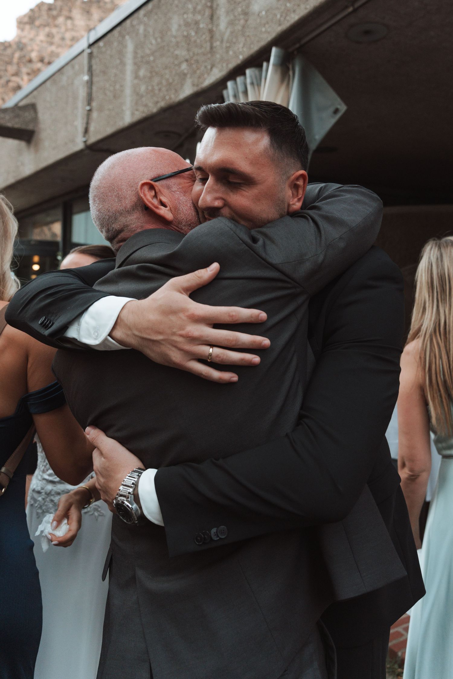 Groom in black suit sharing emotional hug with bald guest at outdoor wedding celebration.
