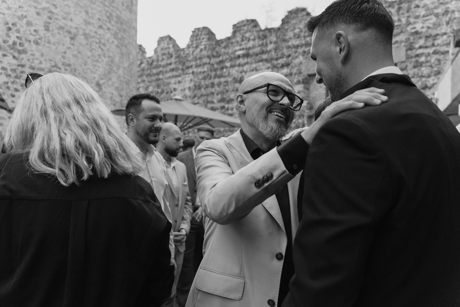 Guests greeting and congratulating groom in white suit at outdoor wedding reception near stone wall.