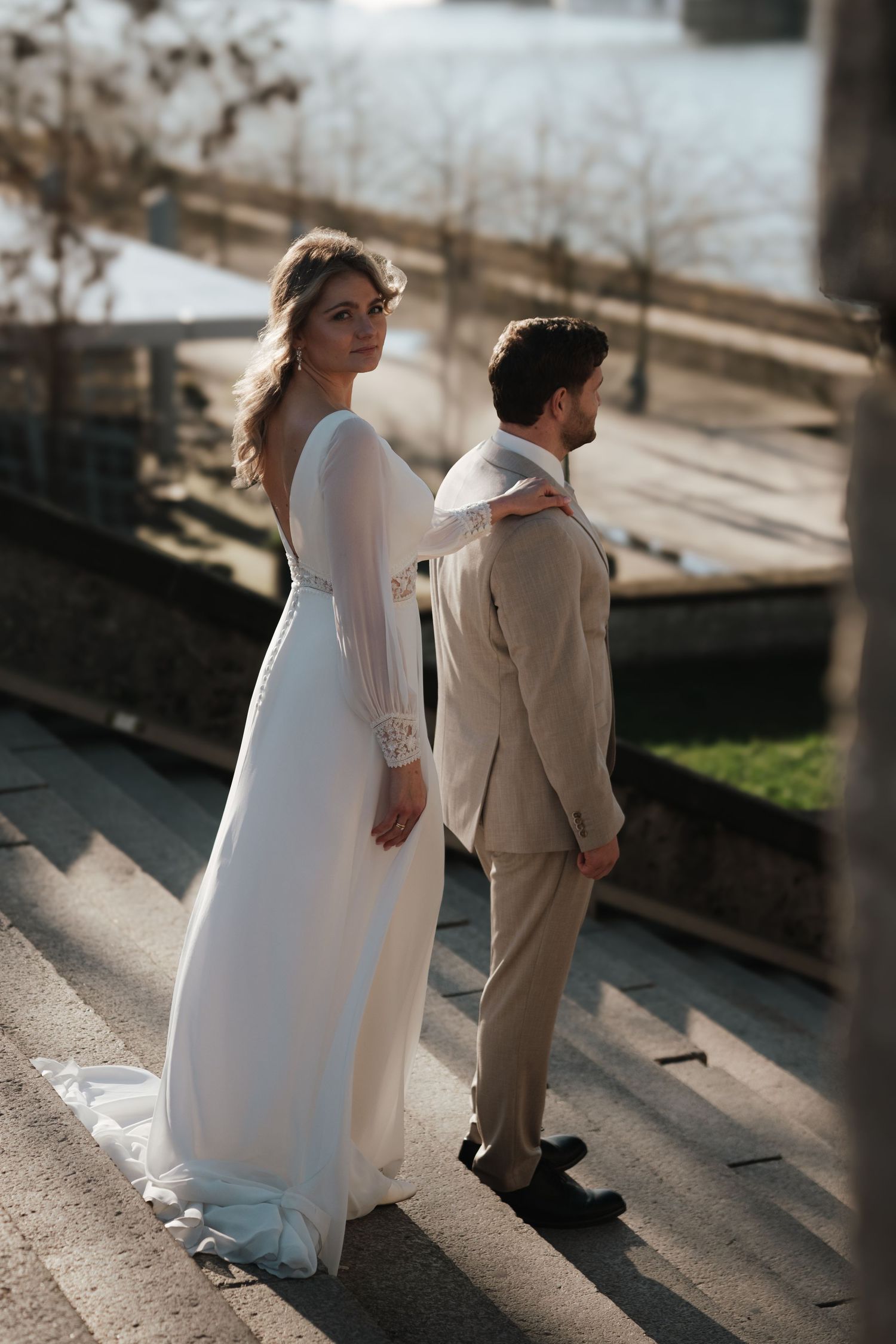Elegant bride in long white gown looks back at camera while holding grooms collar on sunlit riverside steps.