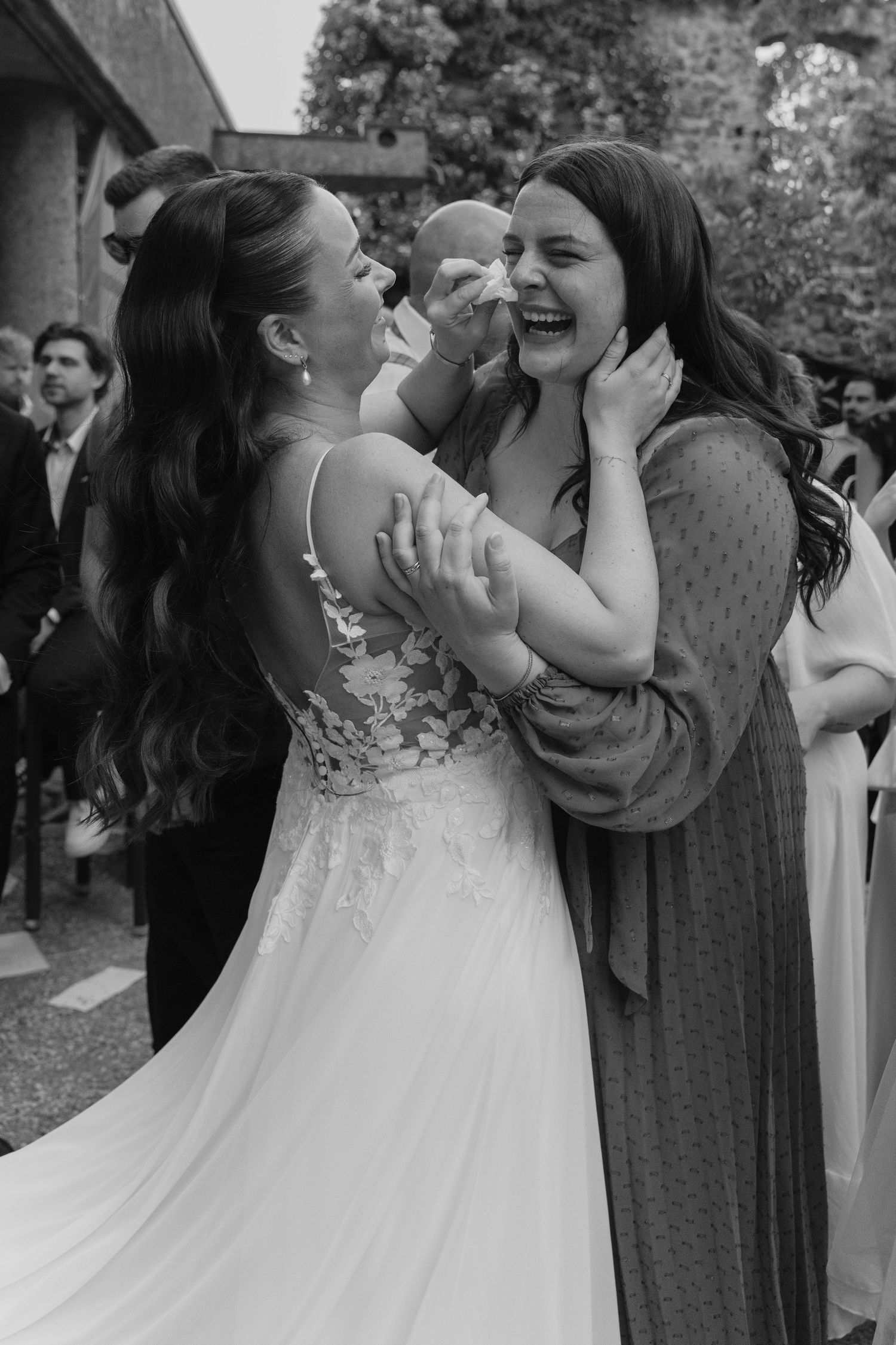 Bride in lace gown laughing joyfully while embracing female guest at outdoor wedding celebration.