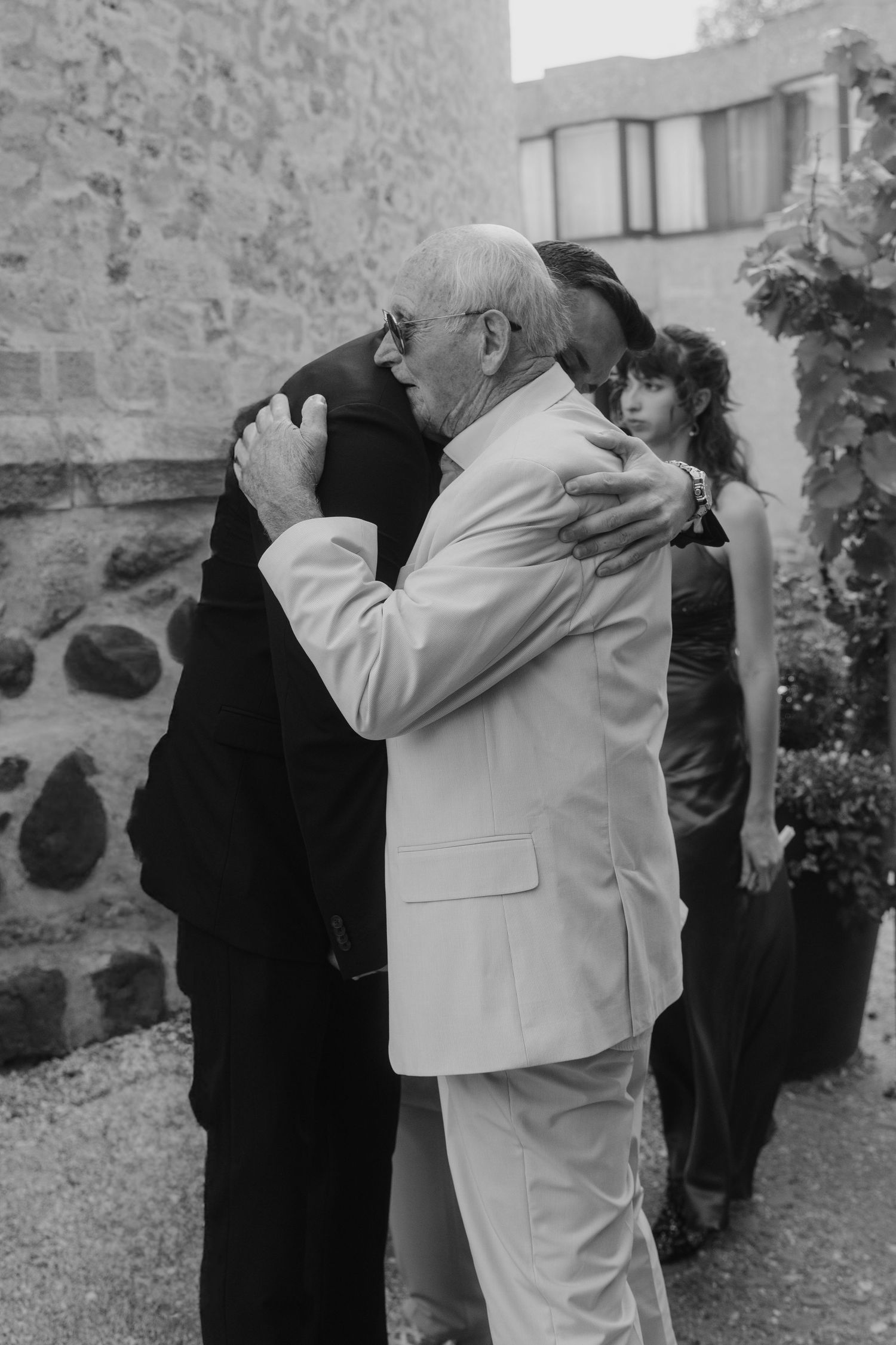 Two elderly men sharing a warm embrace at an outdoor wedding celebration in black and white.