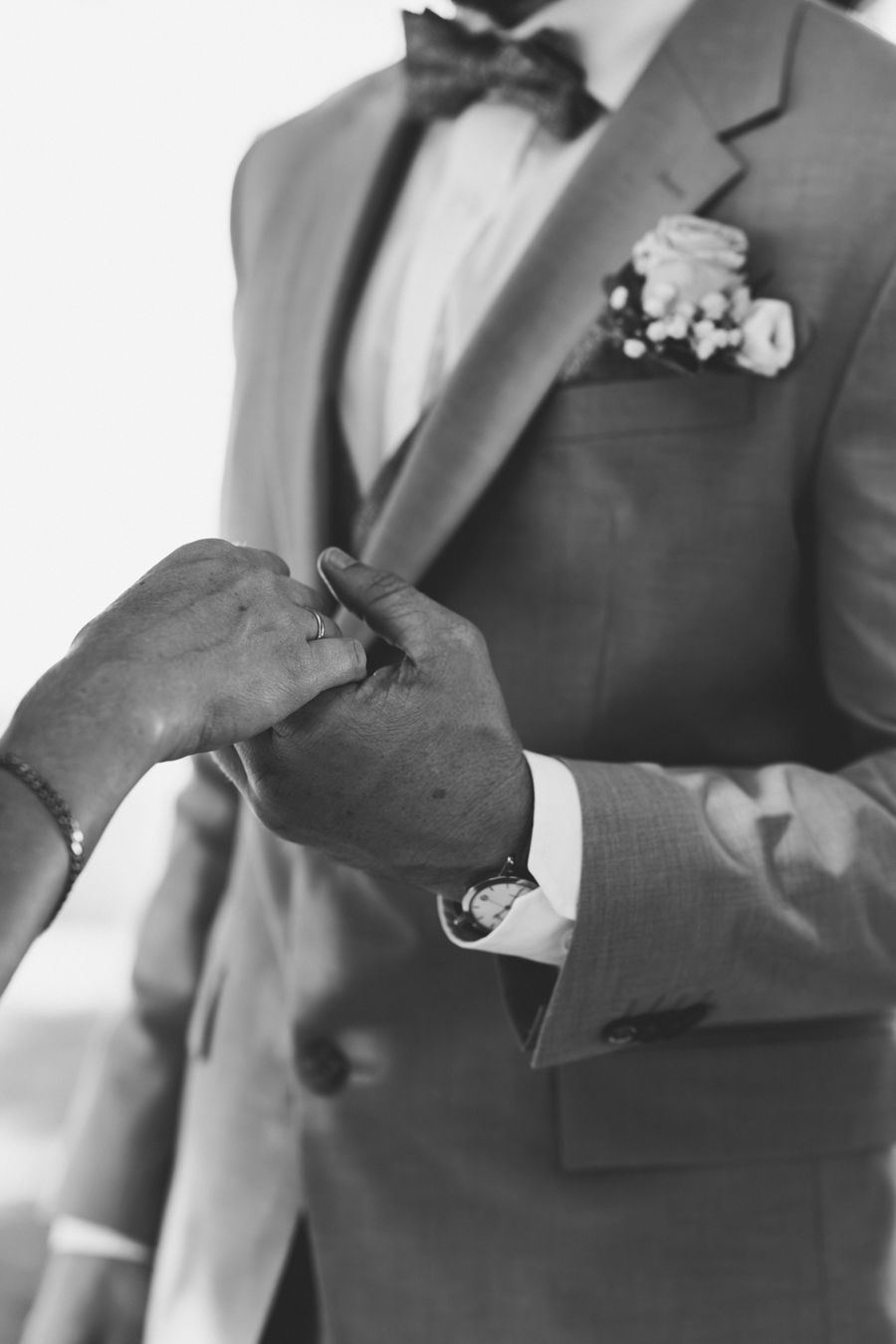 Black and white photo of hands holding groom's lapel in bow tie suit with boutonniere.