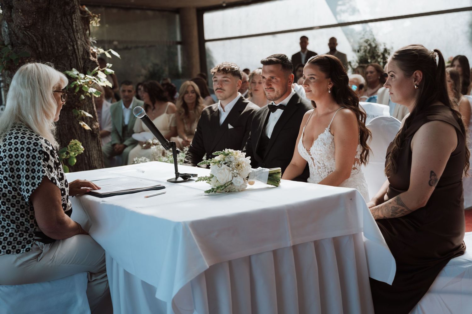 Bride and groom smile at officiant during outdoor wedding ceremony at white draped table with floral bouquet.