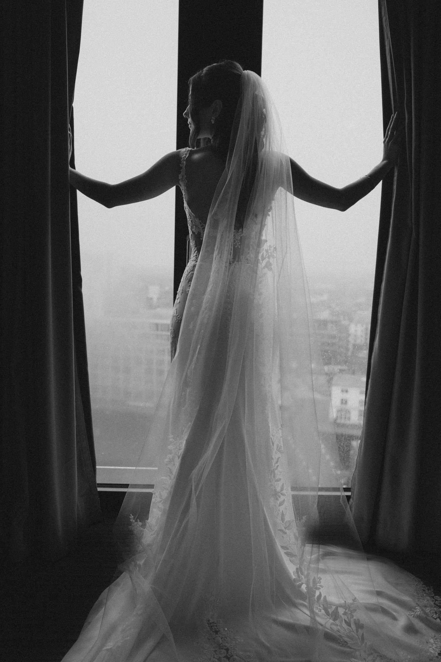 Elegant bride in lace wedding gown and long veil stands silhouetted before large window in black and white photo.