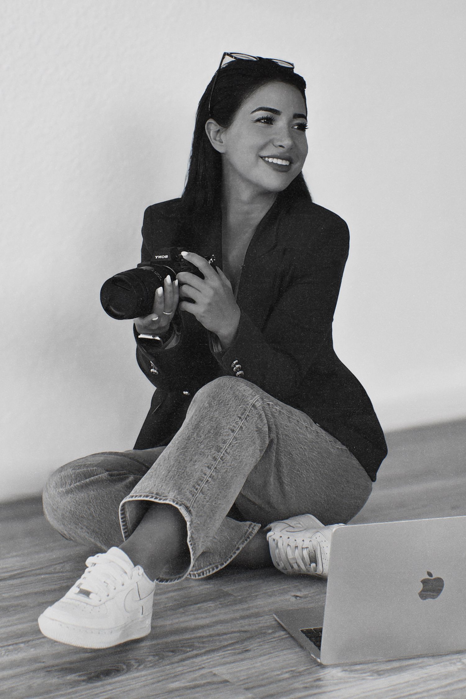 Black and white portrait of a female photographer sitting on floor holding camera with laptop nearby.