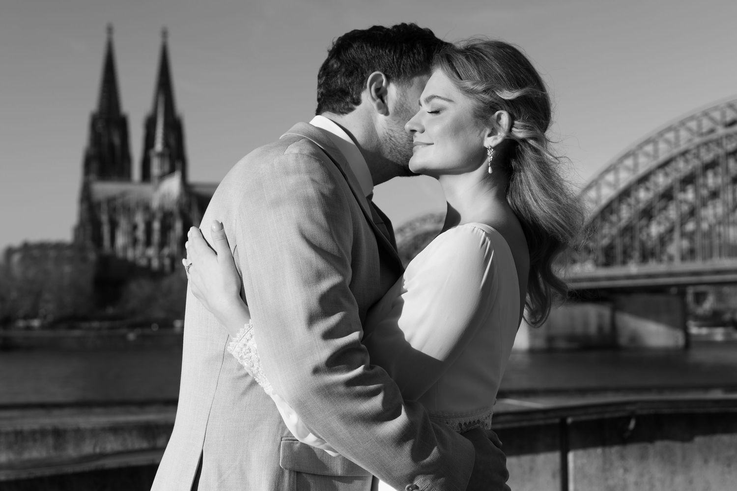 Couple embraces romantically with Cologne Cathedral and Hohenzollern Bridge in background in black and white portrait.