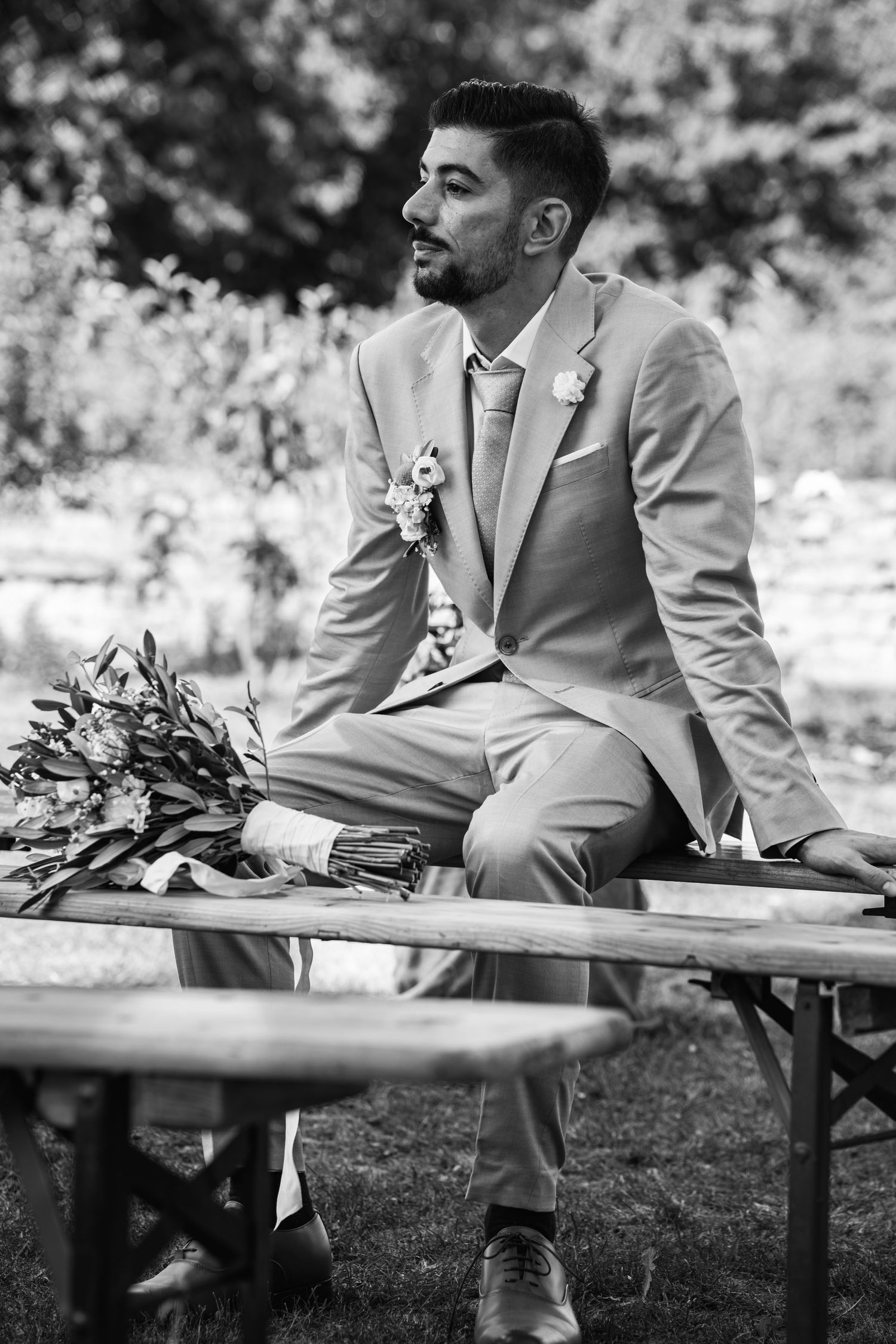 Groom in beige suit sits on bench beside floral bouquet in black and white.