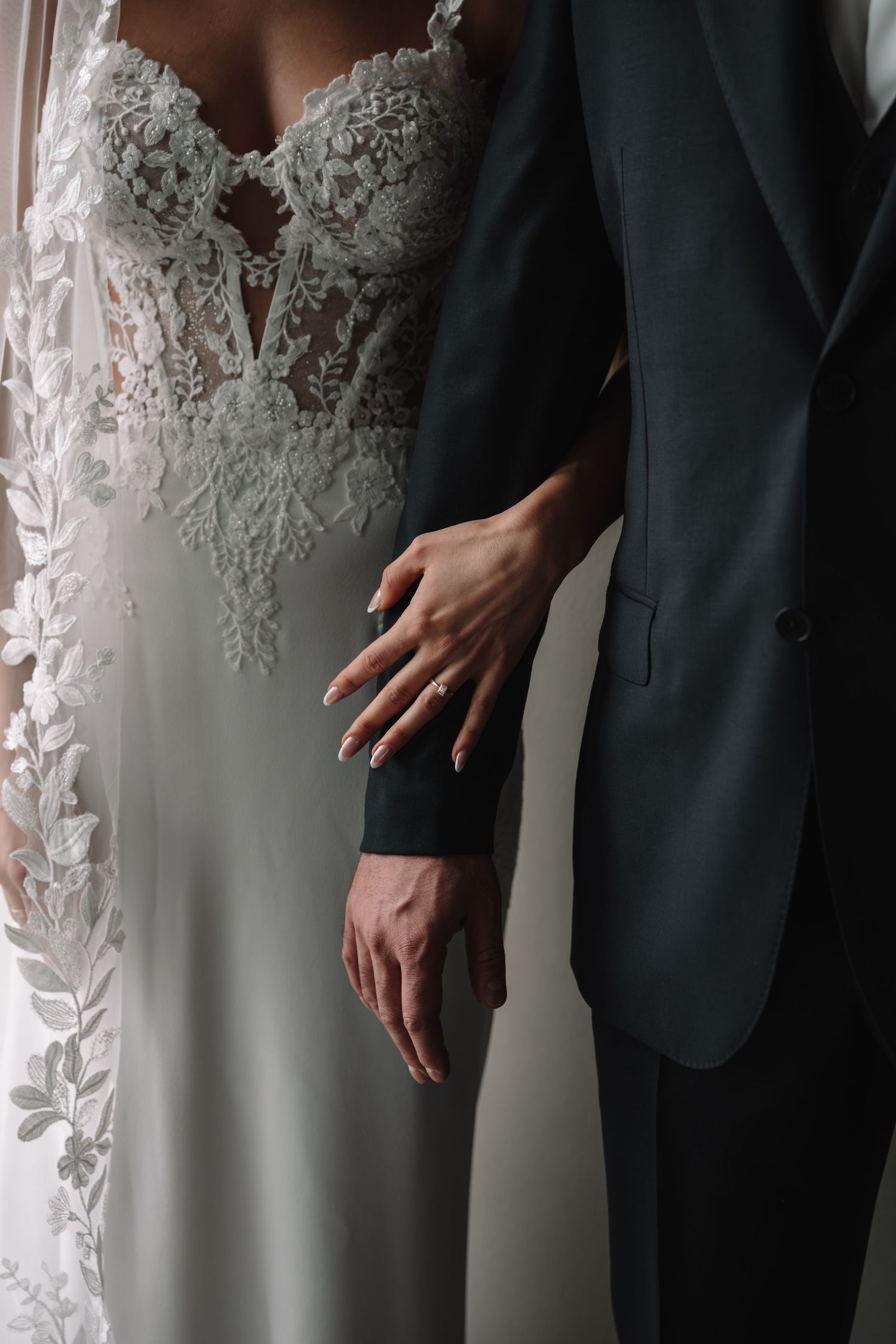 Close-up of bride in lace gown and groom in dark suit holding hands showing wedding ring detail.