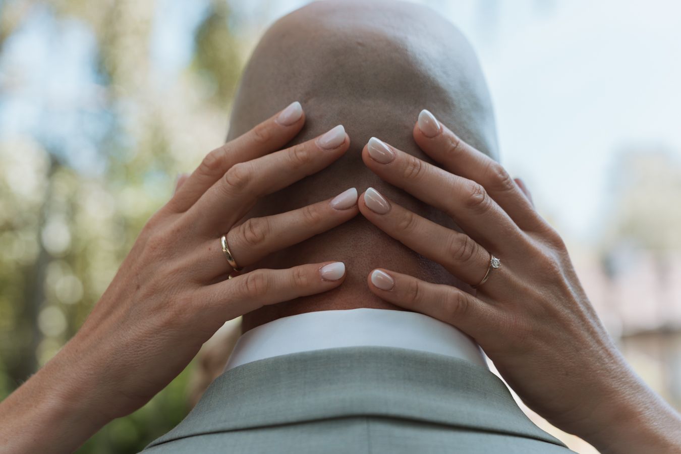 Bride's hands with white manicure and wedding ring resting on groom's head outdoors.