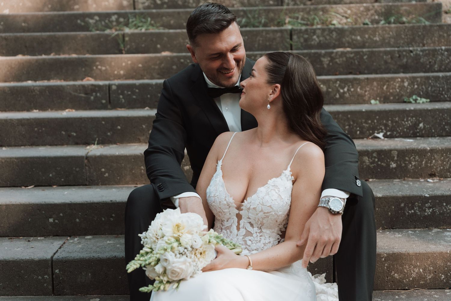 Bride and groom sit on stone steps sharing a romantic moment with white bouquet.