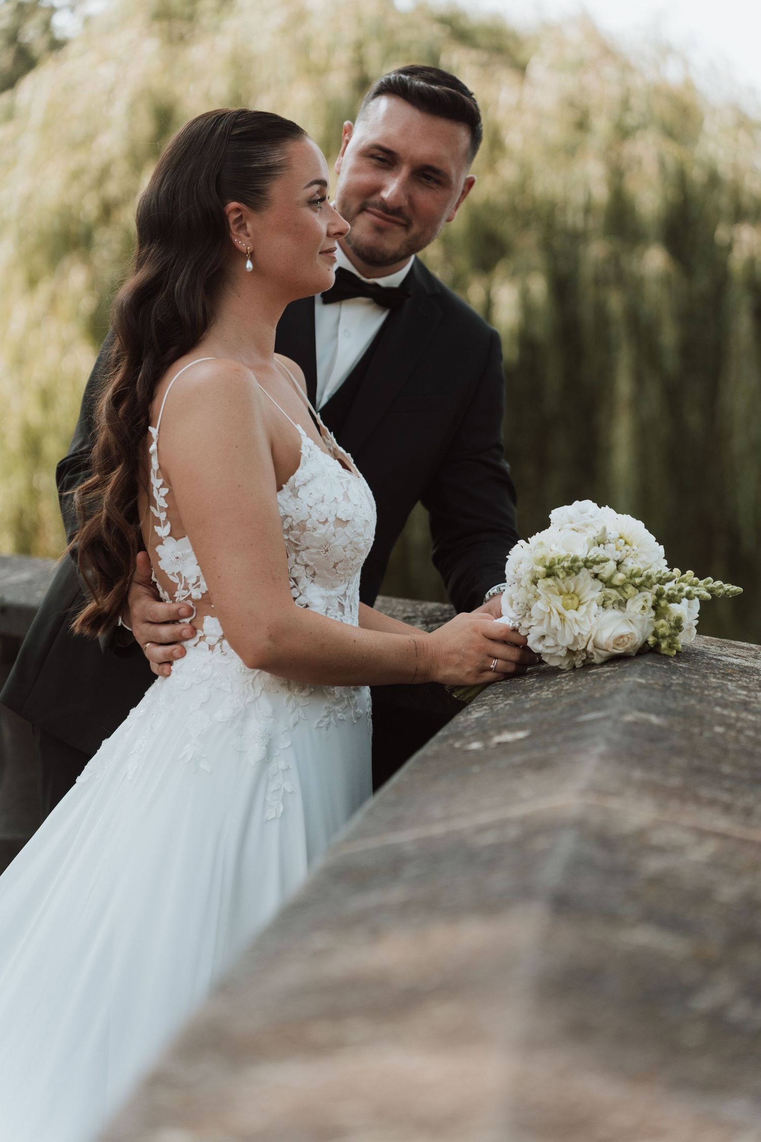 Elegant bride and groom pose together on stone bridge surrounded by lush greenery.