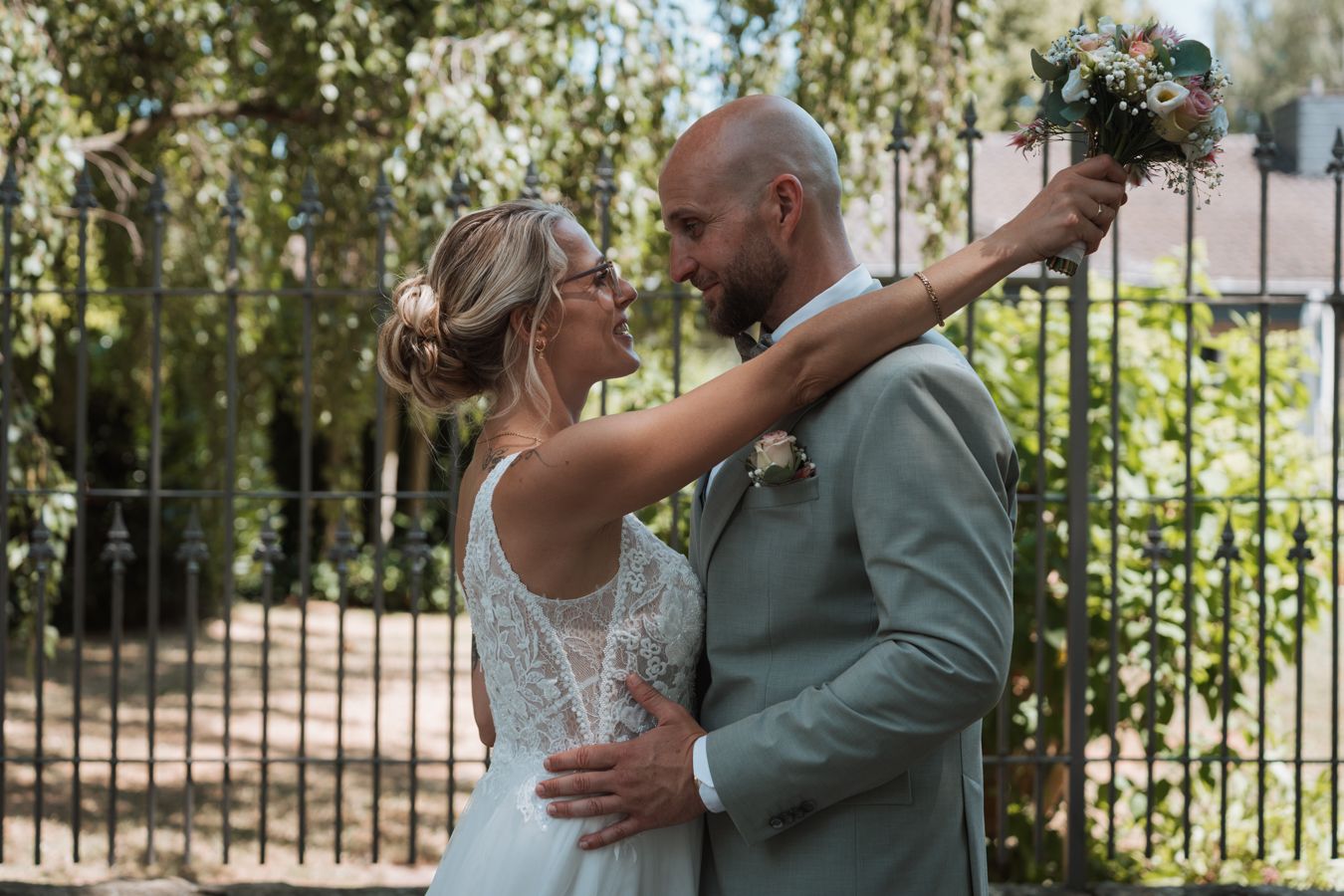 Bride and groom embrace by iron fence as she holds floral bouquet overhead.