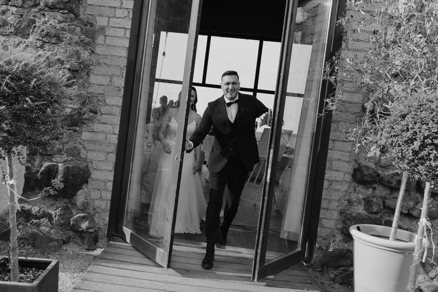 Excited groom in black tuxedo rushes through glass doors with bride visible in reflection behind.