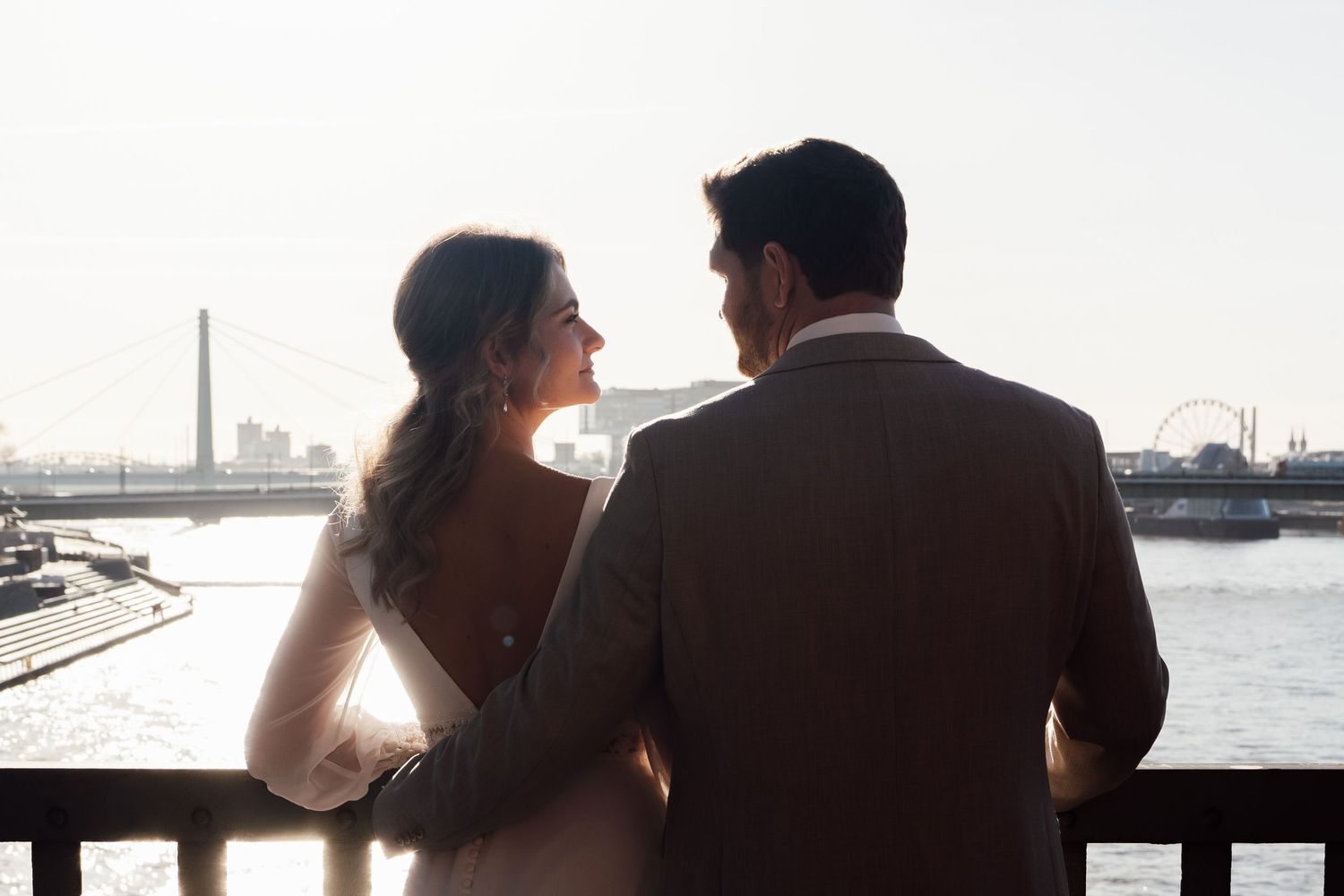 Bride and groom share a quiet moment overlooking a sunlit waterfront skyline from a wooden railing.
