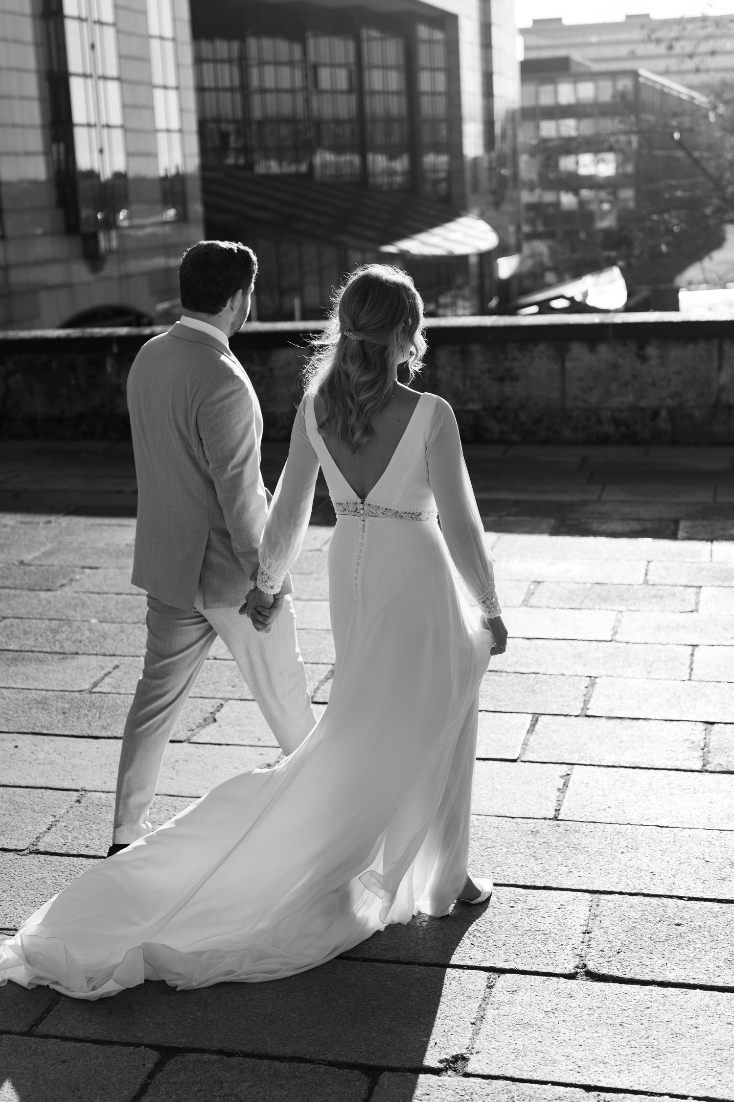 Black and white photo of bride and groom holding hands walking on cobblestone street in elegant wedding attire.
