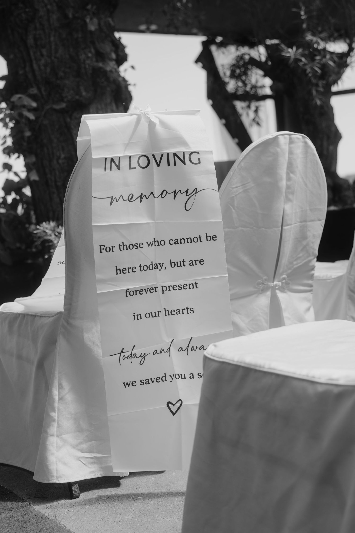 Black and white photo of white wedding chair with In Loving Memory sign honoring absent loved ones.