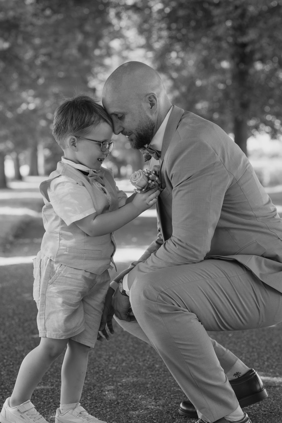 Bald bearded groom in tan suit shares tender nose-to-nose moment with young boy in black and white photo.