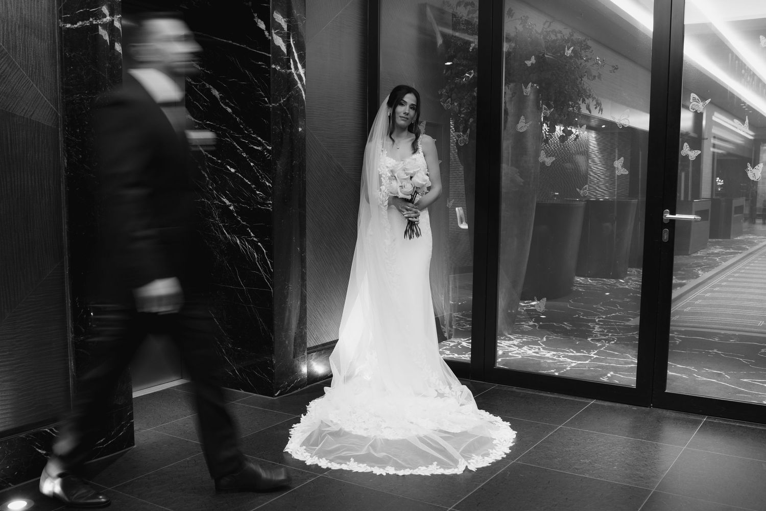 Bride in white gown and veil stands poised at venue entrance as blurred figure walks past in black and white.