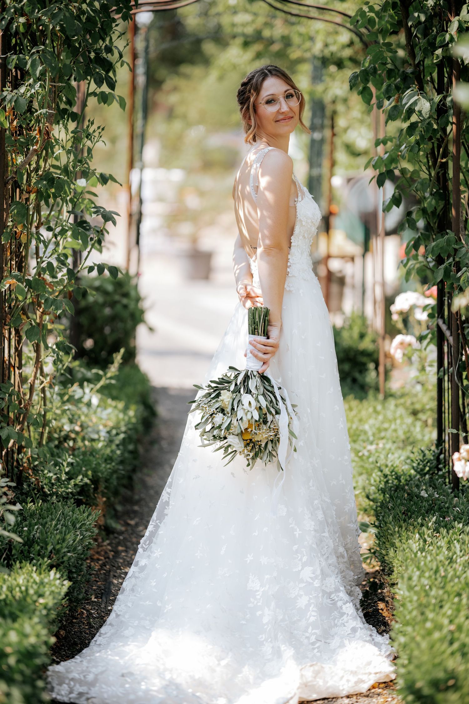 Beautiful bride in a floral lace wedding dress holding a bouquet in a lush garden archway.