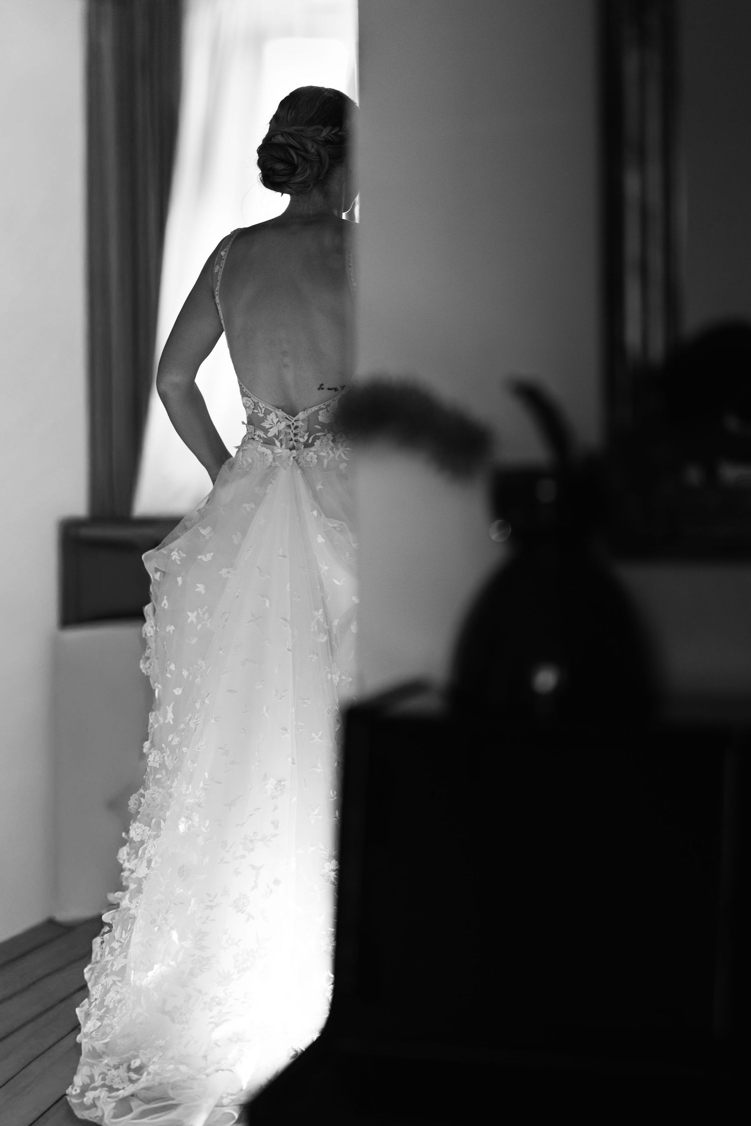 Black and white photo of bride in backless embellished wedding gown with ruffled train standing by window.