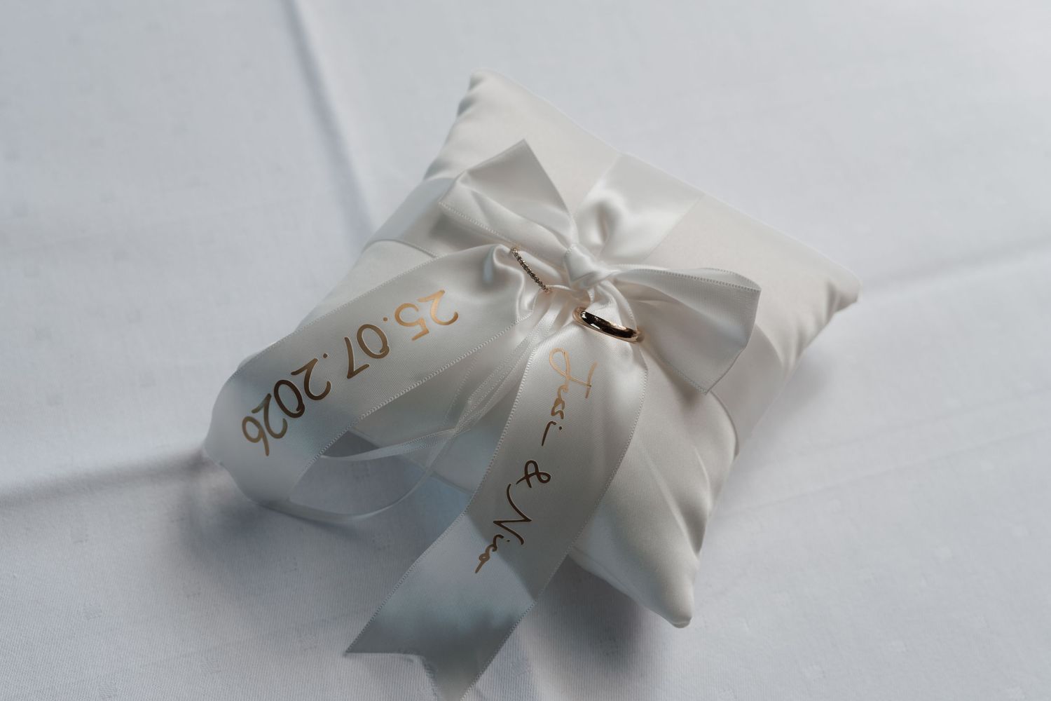White satin wedding ring pillow with bow, dated 25.07.2020, holding gold wedding bands.
