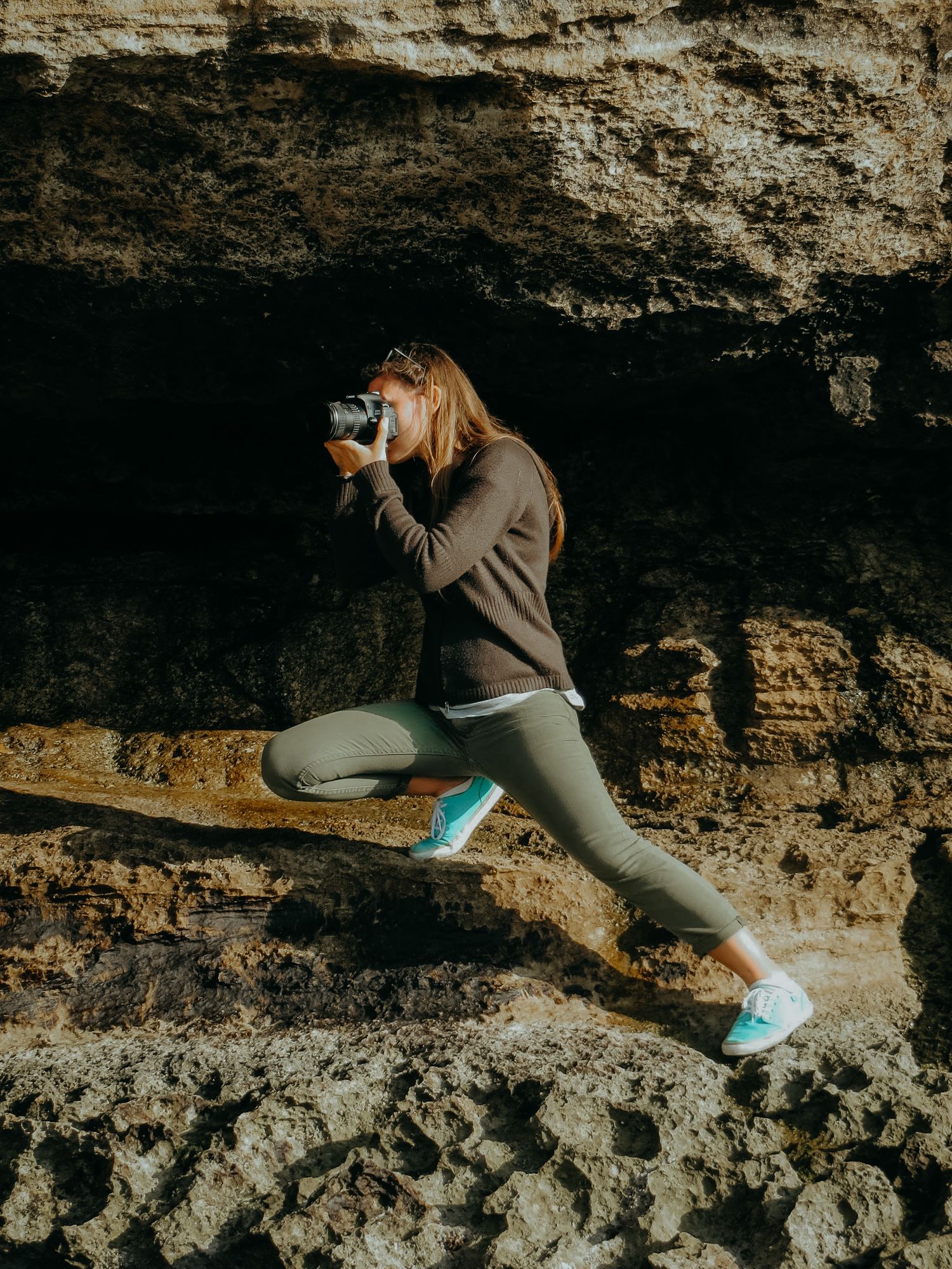 A woman in green leggings photographs rocky coastal cliffs while kneeling on layered stone formations.