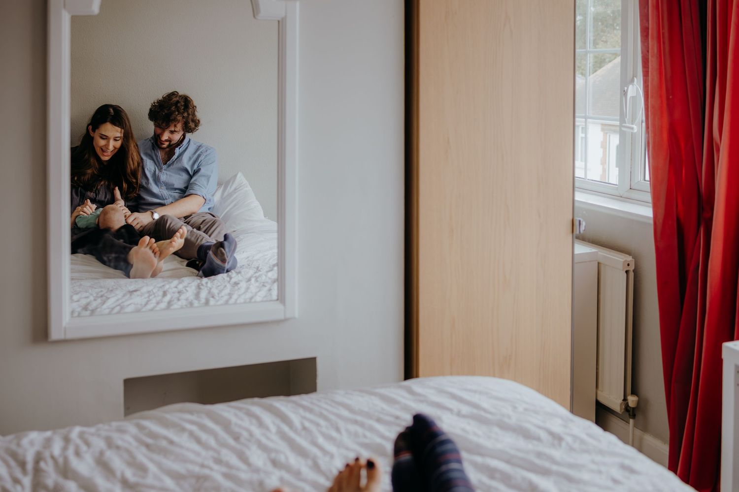 Couple sitting on bed with newborn baby reflected in bedroom mirror.