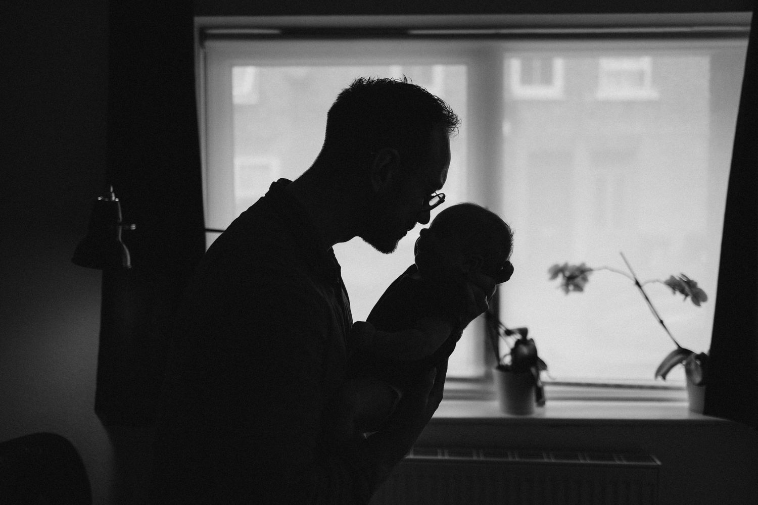 Black and white silhouette of a father kissing his newborn baby by a window.