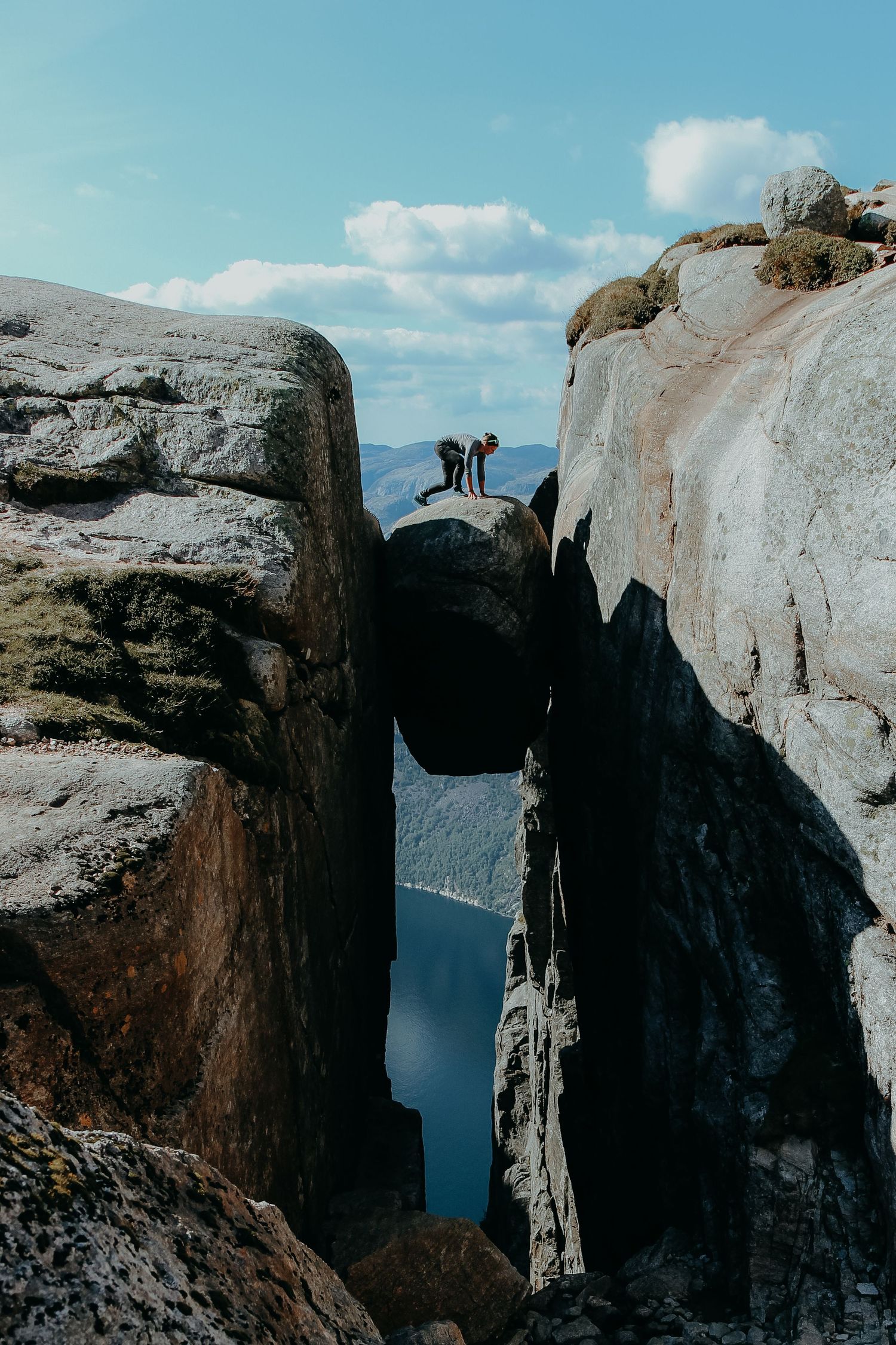Two people stand on the famous Kjeragbolten boulder wedged between rocky cliffs above a Norwegian fjord.