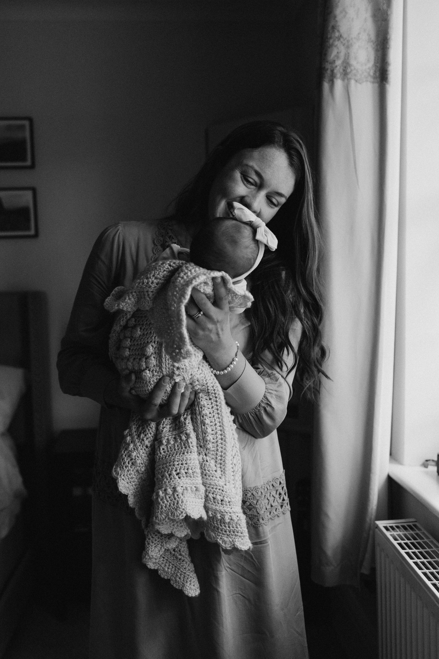 Black and white photo of a mother kissing her newborn baby by a window.