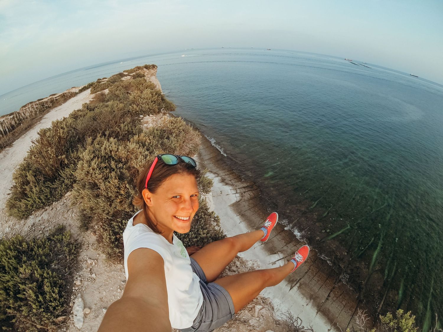 A smiling girl takes a selfie on a rocky coastal cliff with turquoise ocean water stretching behind her.