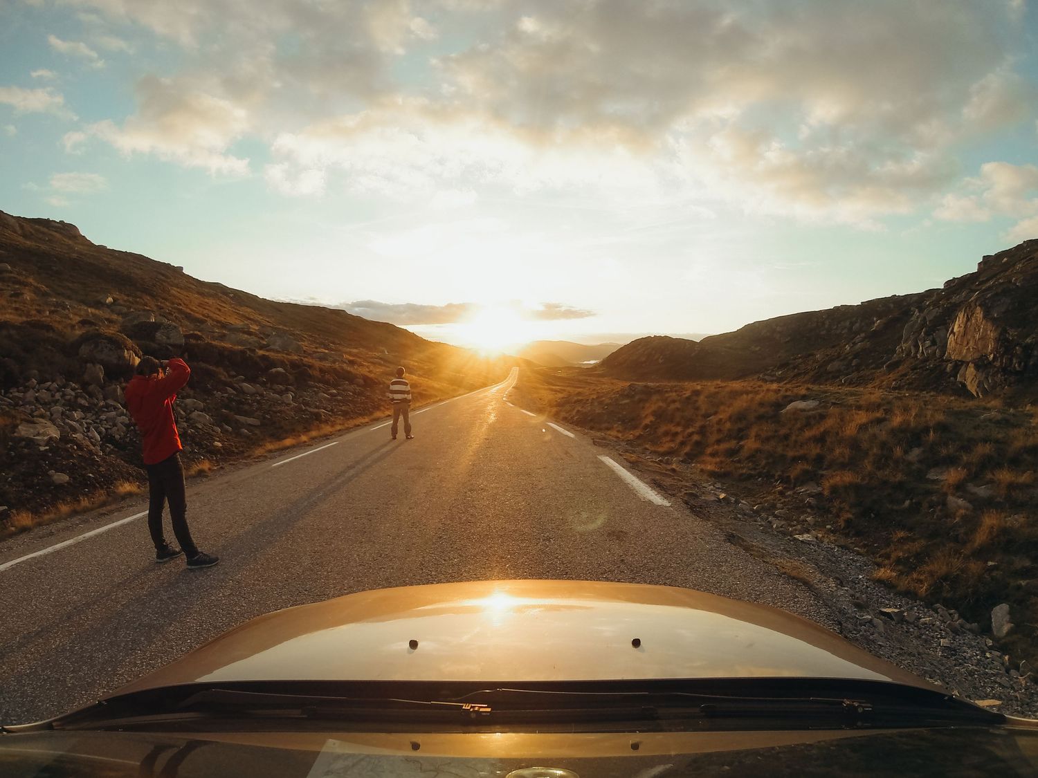 A person stands on a scenic road at sunset, viewed from a car hood, with golden hills on both sides.